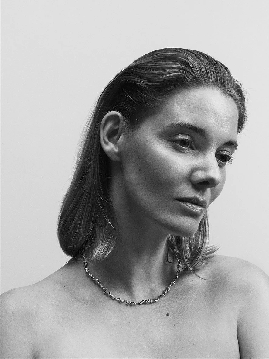 Black-and-white portrait of a woman with shoulder-length hair, wearing a necklace, looking down and to the side against a plain background. Pernille Lauridsen Jewellery campaign. Model waring lovely jewellery and in a soft light setting.