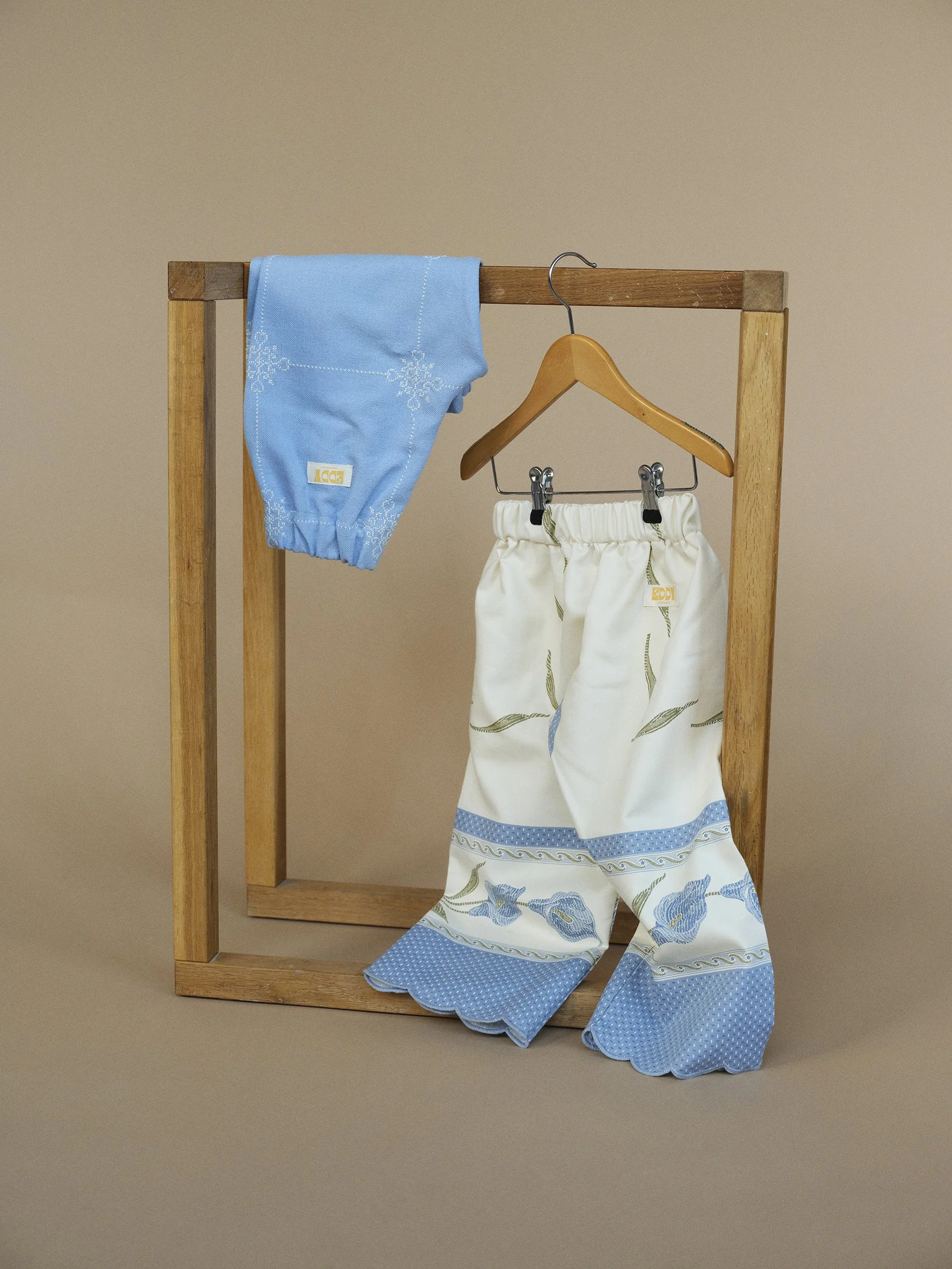 Baby clothes hanging on a wooden stand, including white pants with a blue floral pattern and a blue garment draped over the top.