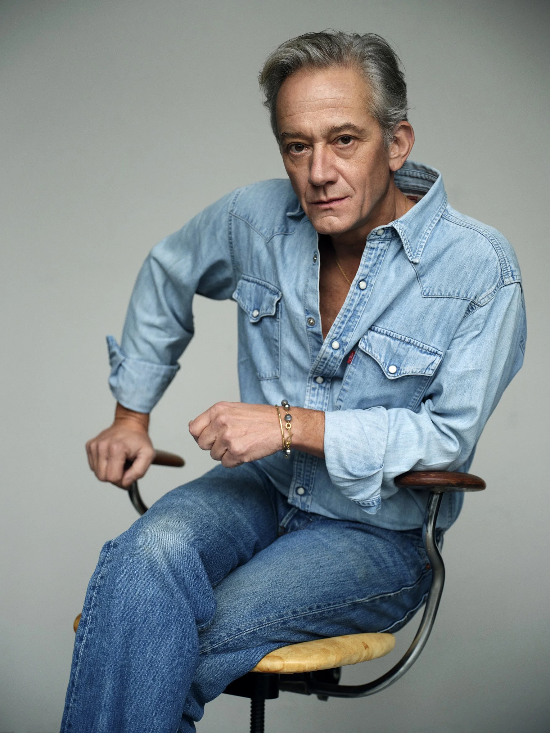 A mature man with gray hair wearing a light denim shirt and jeans, sitting on a wooden and metal chair, looking directly at the camera with a serious expression.