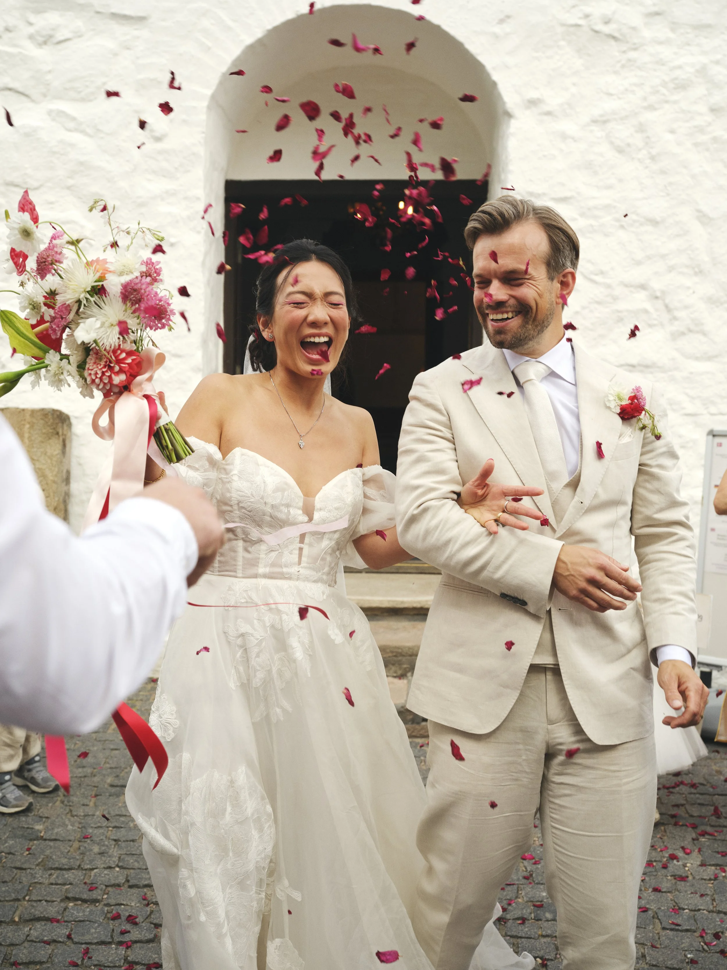 A bride and groom celebrating their wedding with rose petals falling around them outside a building.