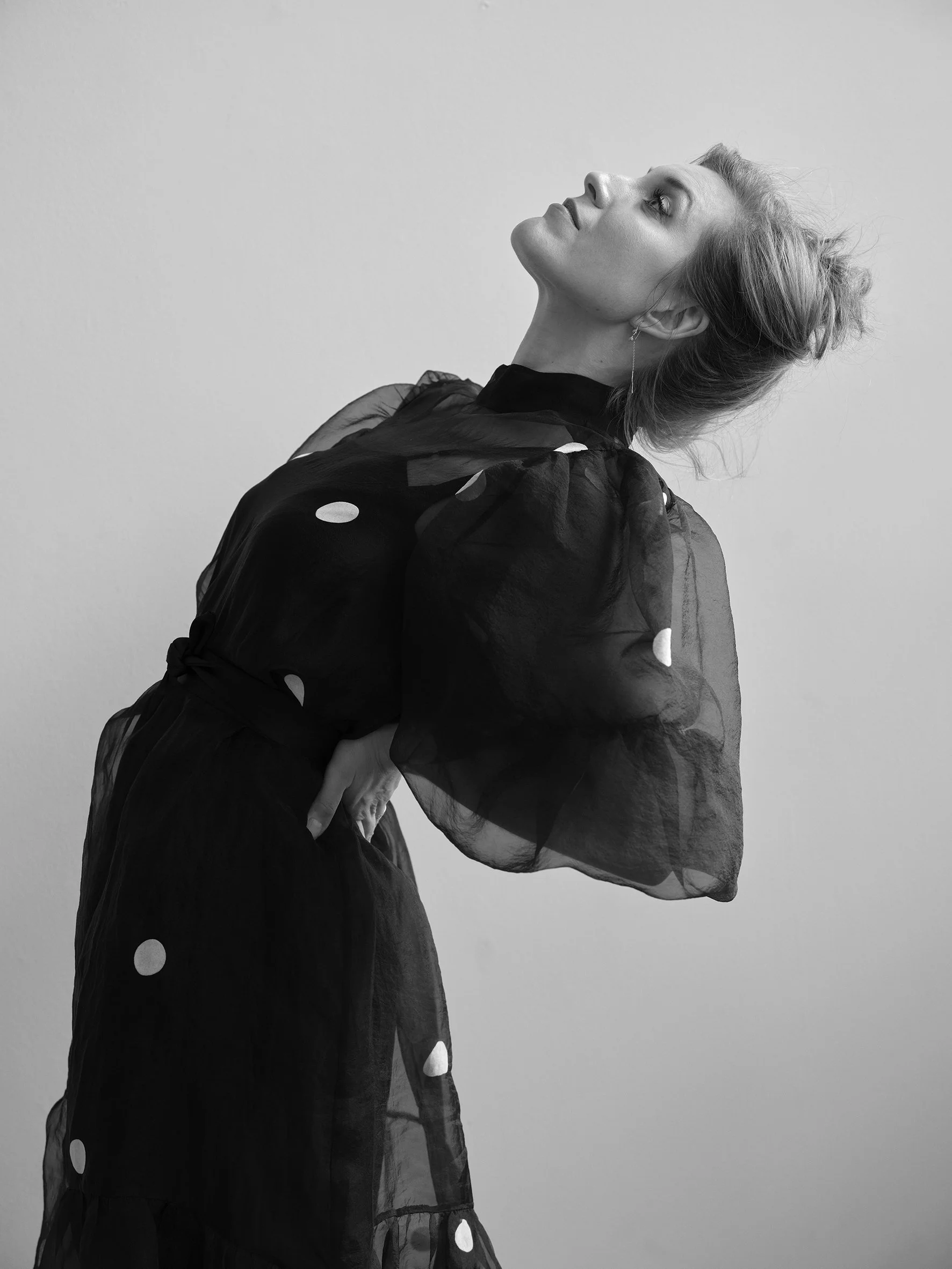 Black and white photo of a woman with her head tilted back, wearing a sheer dress with polka dots and her hand on her hip.
