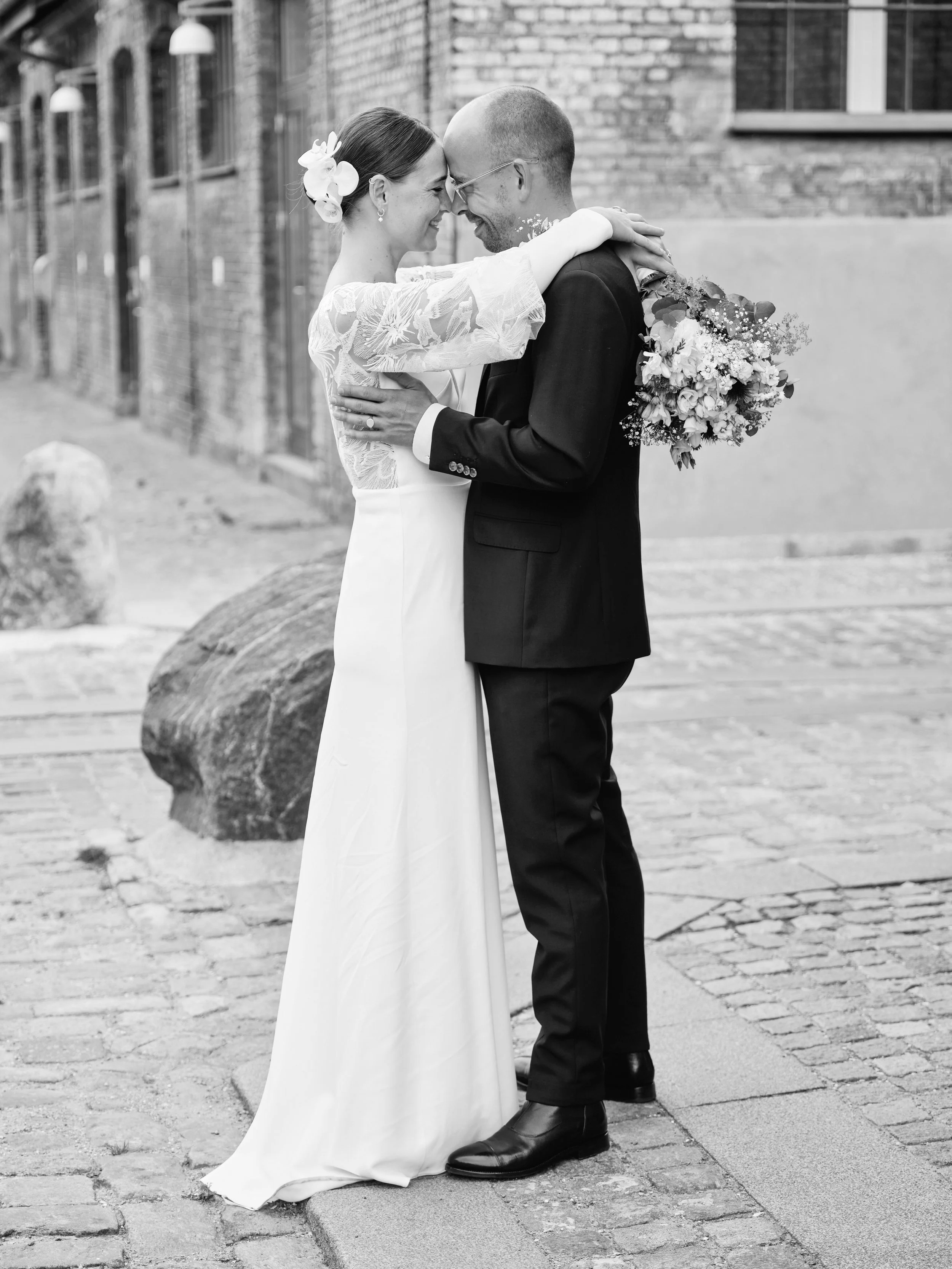 A black-and-white photo of a bride and groom embracing each other, with foreheads touching, outdoors on a cobblestone street. The bride is wearing a long white dress with lace sleeves and has flowers in her hair. The groom is in a dark suit and glasses. The bride is holding a bouquet of flowers behind the groom's back.