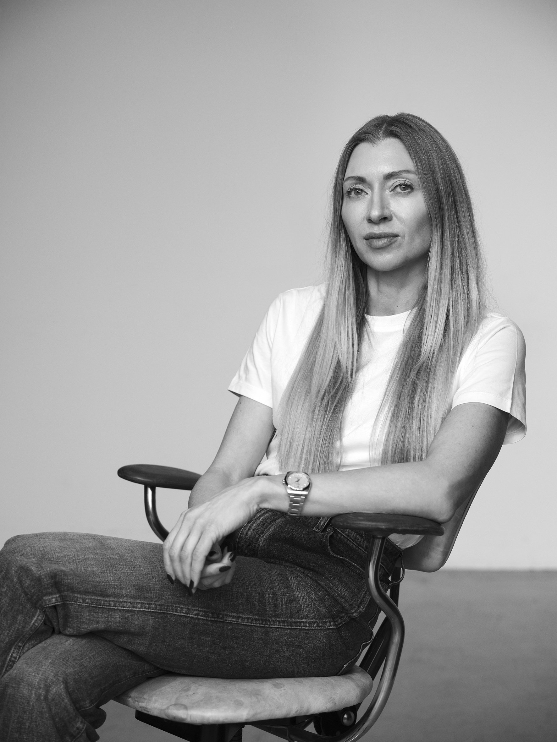 Black and white portrait of a woman with long straight hair, sitting on a chair with her arms crossed, wearing a white t-shirt, jeans, and a watch, against a plain background.