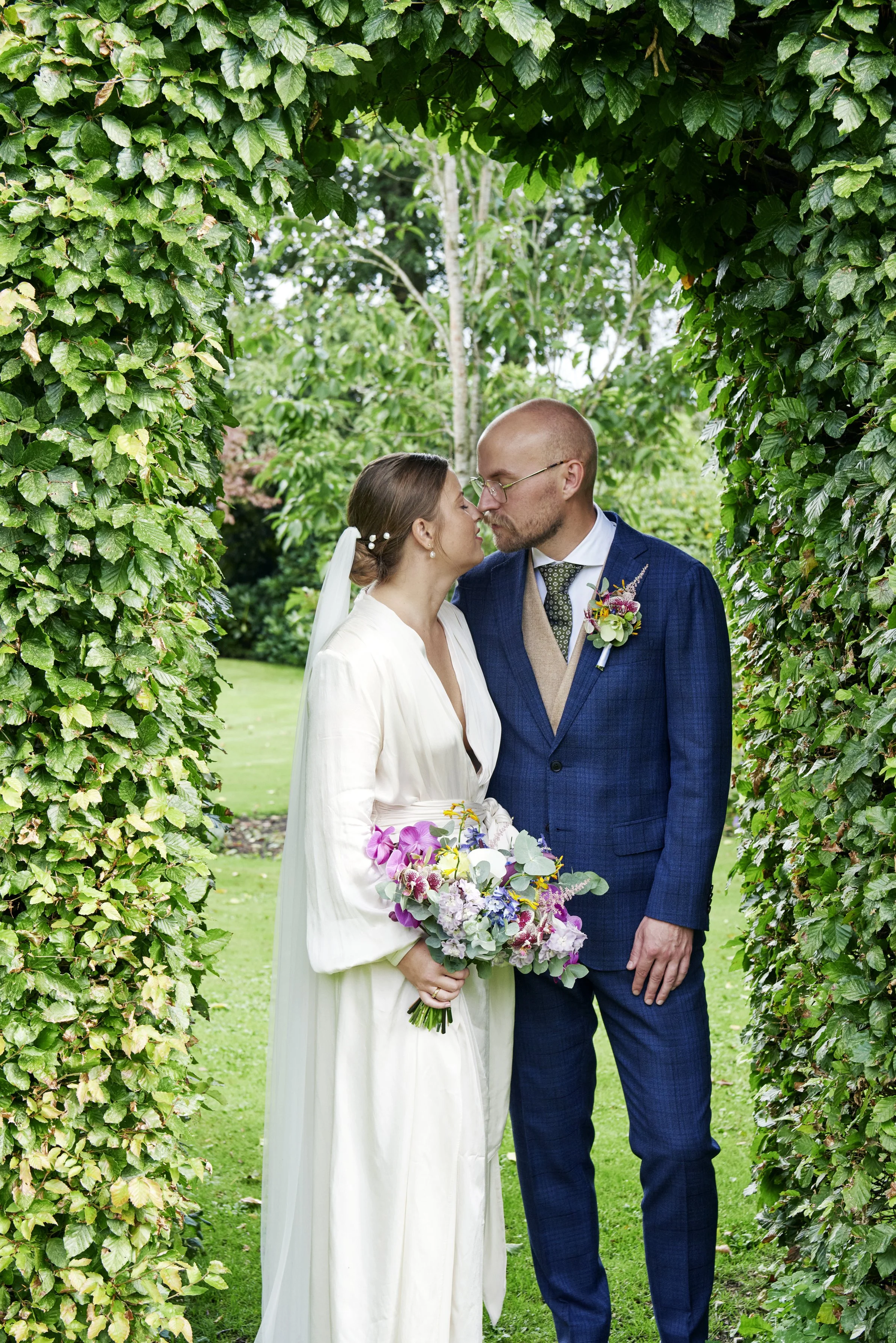 A bride and groom stand close with foreheads touching, under a green archway made of leaves, in a garden.