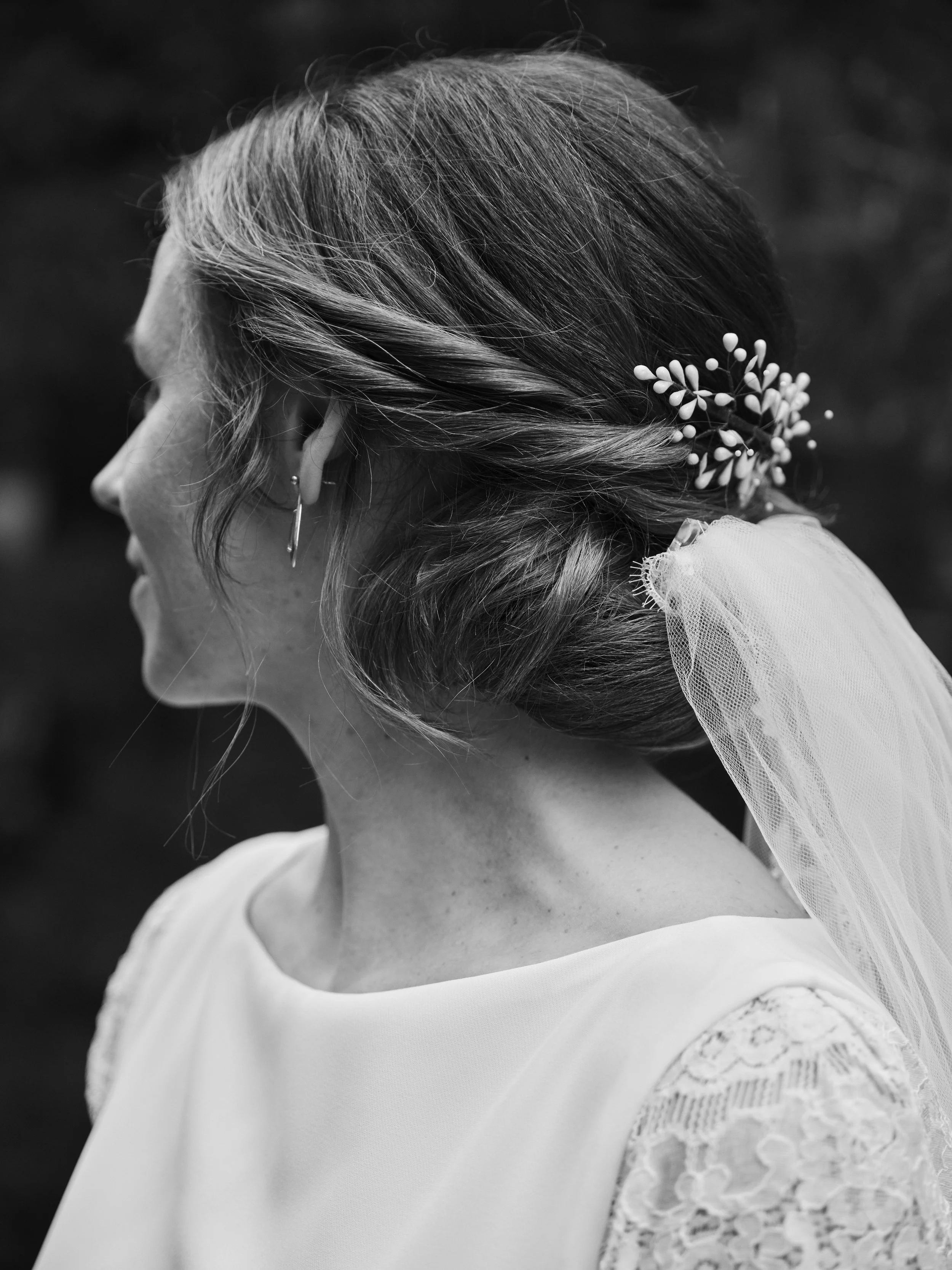 A woman in profile wearing a wedding dress with lace sleeves, her hair styled in an elegant updo adorned with a small floral hair accessory, and a veil attached at the back.