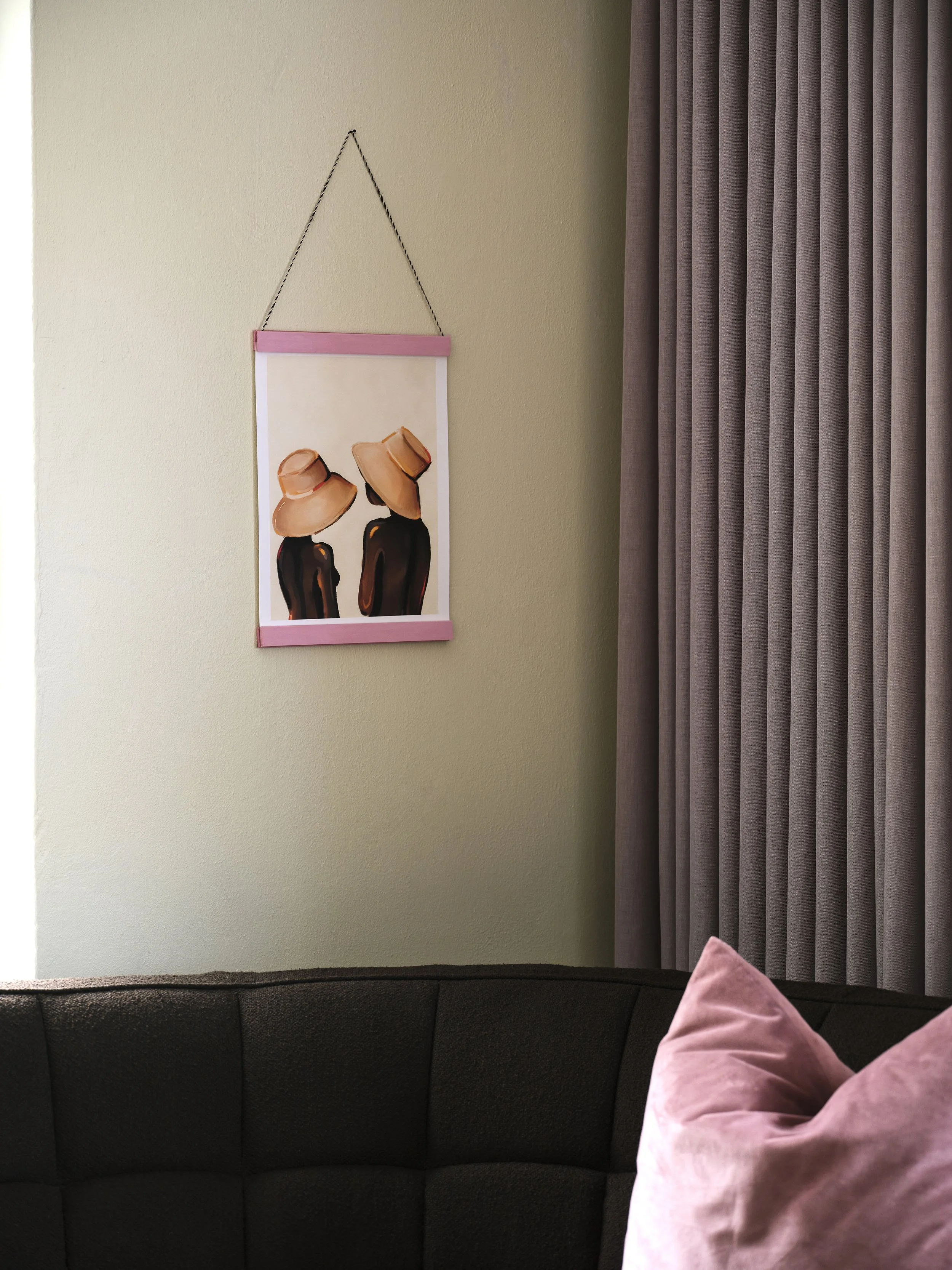 Living room corner with a framed artwork of two people wearing large hats on a beige wall, paired with a black couch and pink velvet pillows, and grey curtains.