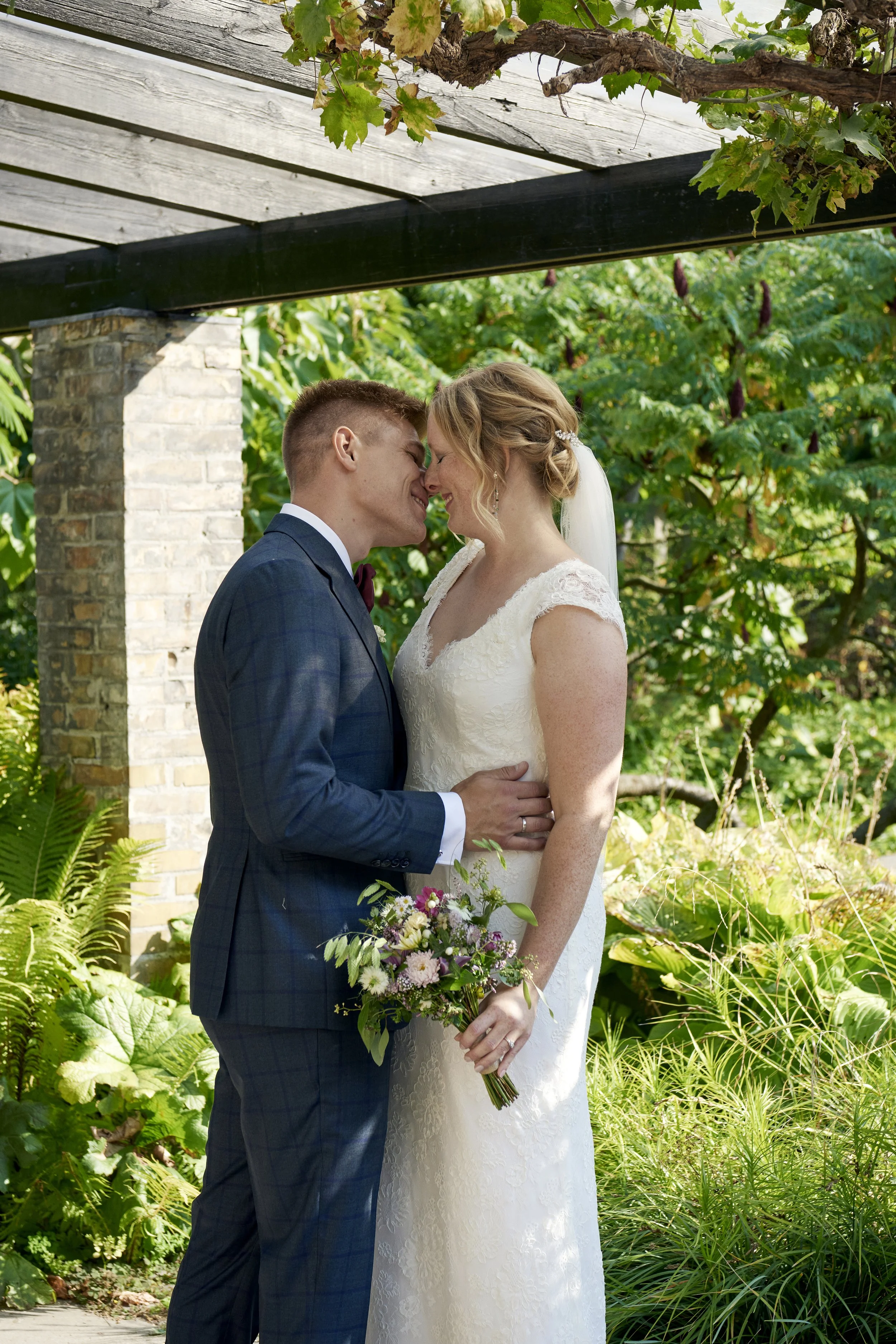 A newlywed couple standing close together under a garden pergola, nose to nose, holding each other with a bouquet of flowers in hand, surrounded by lush greenery.