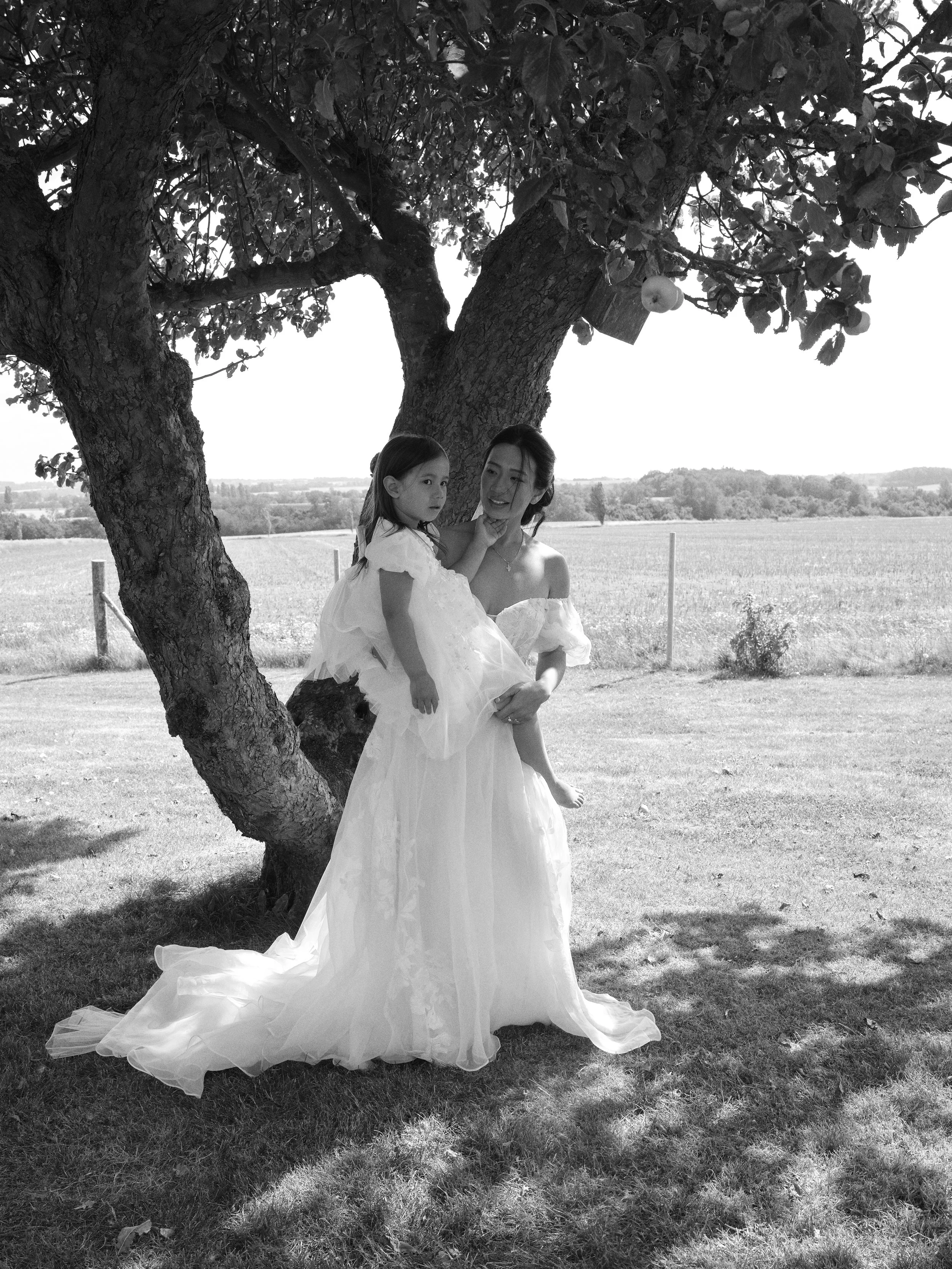A woman in a wedding dress holding a young girl in a white dress under a large tree in an open field.
