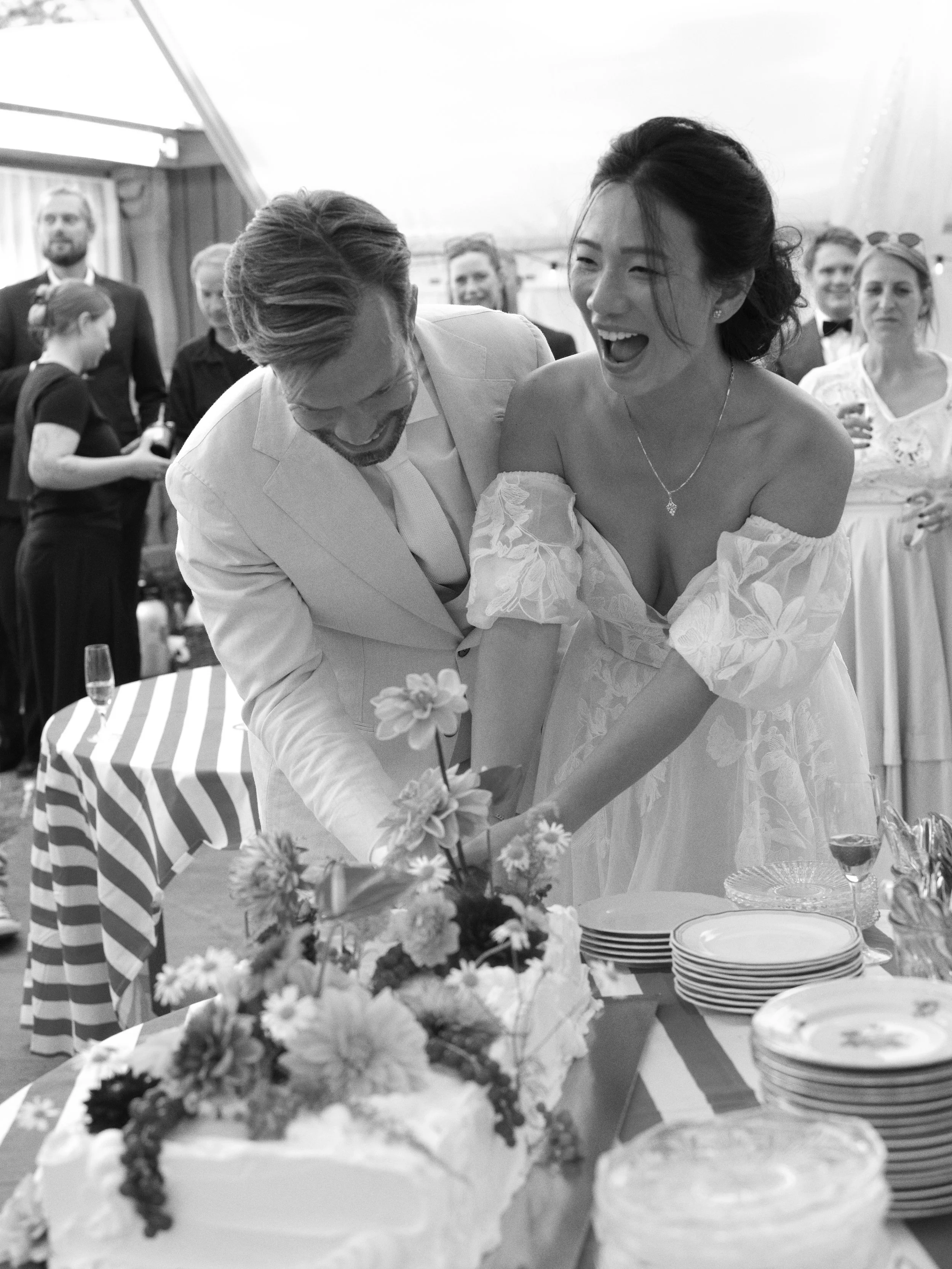 A bride and groom cut a wedding cake together at a wedding reception with guests watching and celebrating.