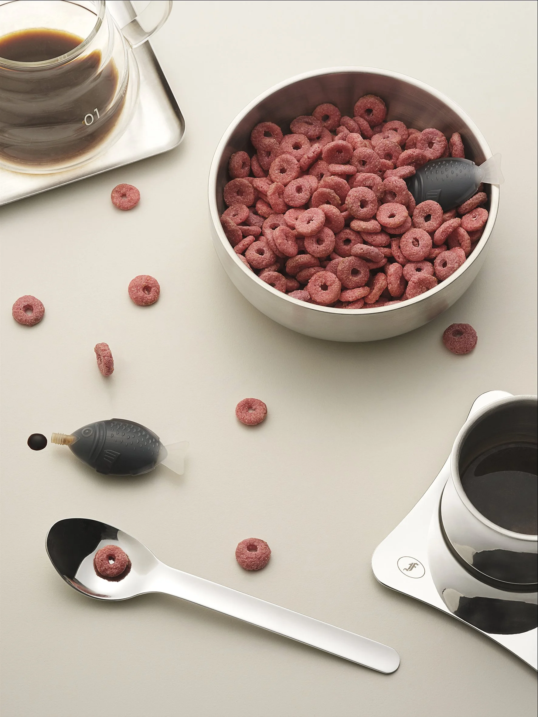 A bowl of pink cereal with fish-shaped toys, a spoon, a glass of black coffee, and a jar of black liquid on a white surface.