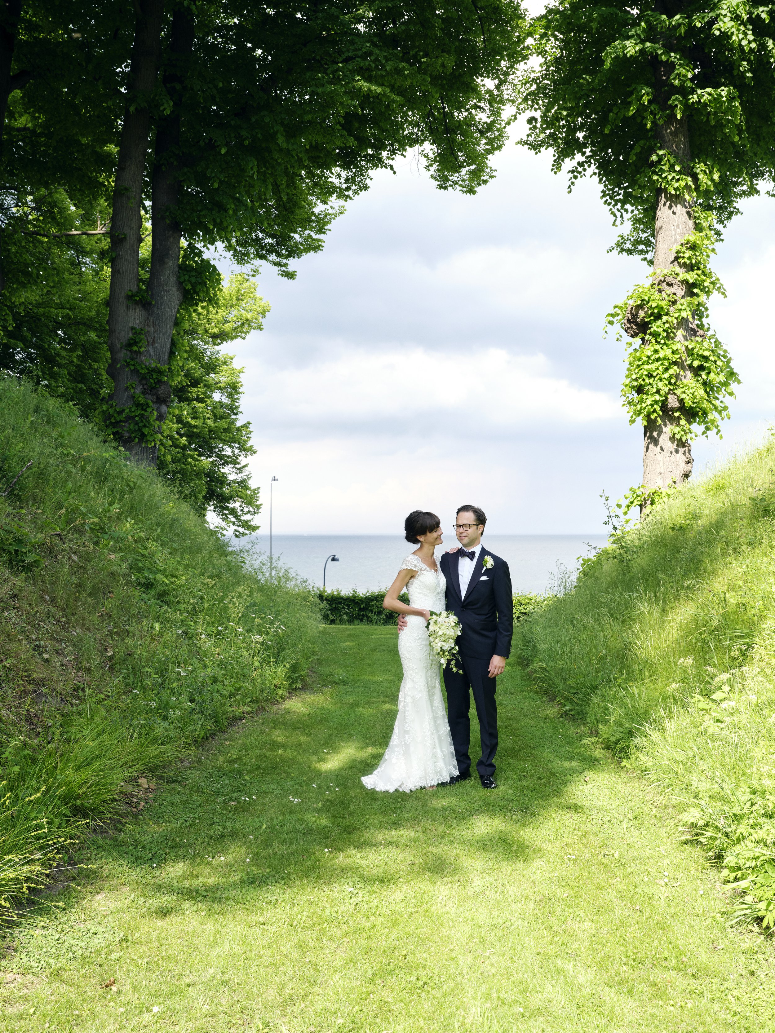 A bride and groom in wedding attire standing together on a grassy path under trees with the ocean in the background.