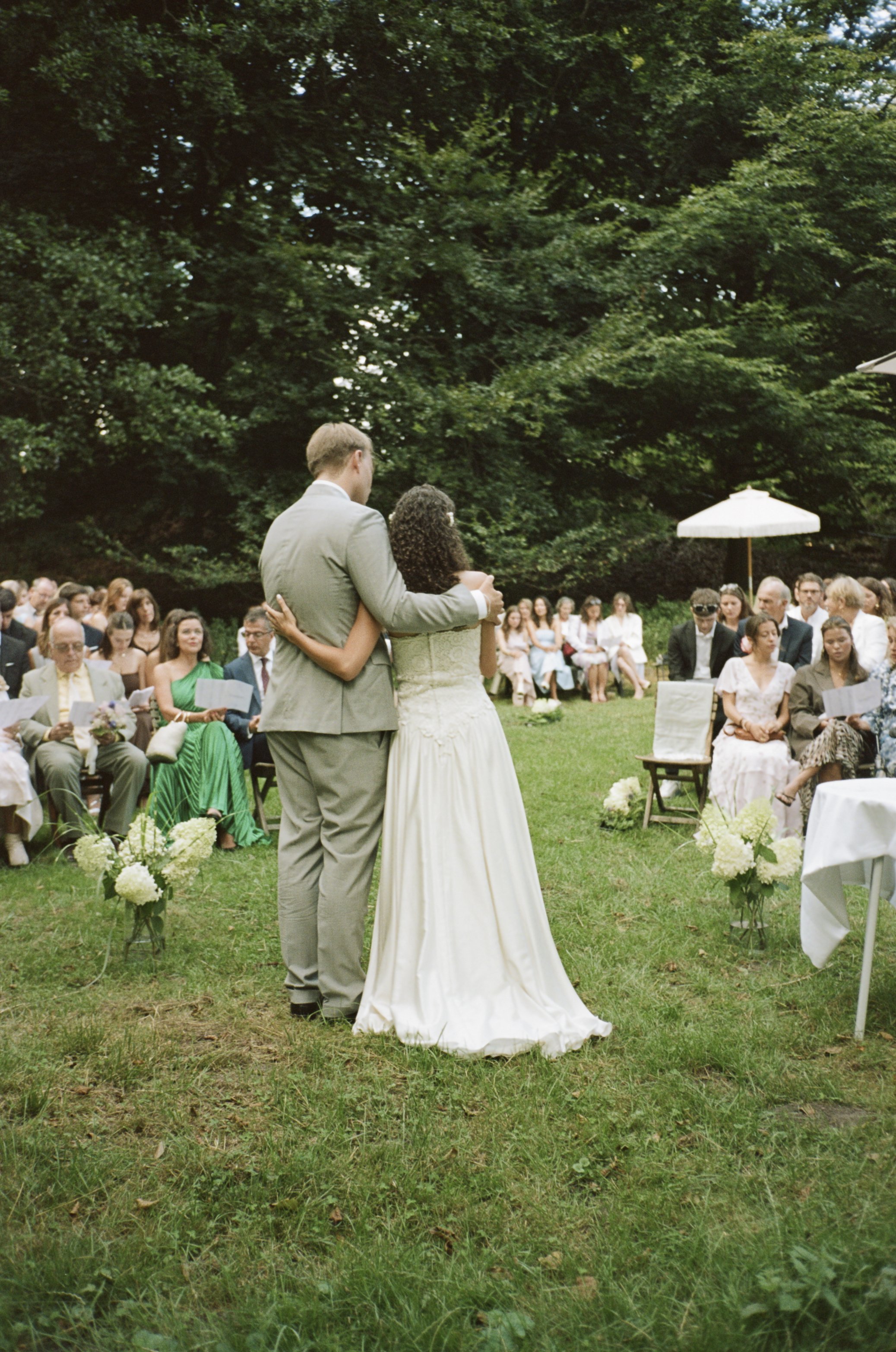 A bride and groom standing close together during an outdoor wedding ceremony. The groom is dressed in a light gray suit, and the bride is wearing a white gown. They are surrounded by seated guests on a grassy area with trees in the background.