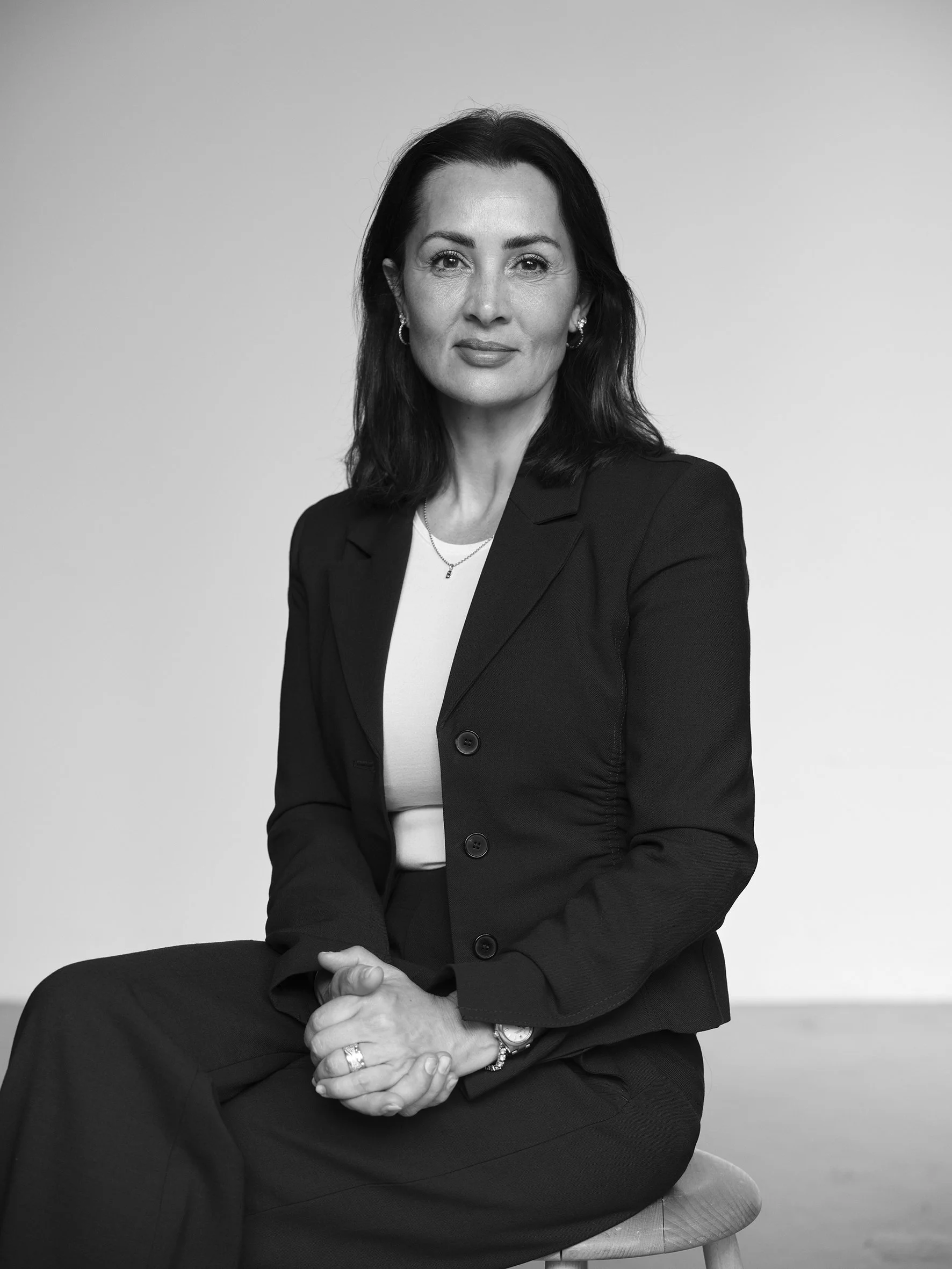 Black and white portrait of a woman with dark hair, wearing a black blazer and white top, sitting on a stool against a plain background.