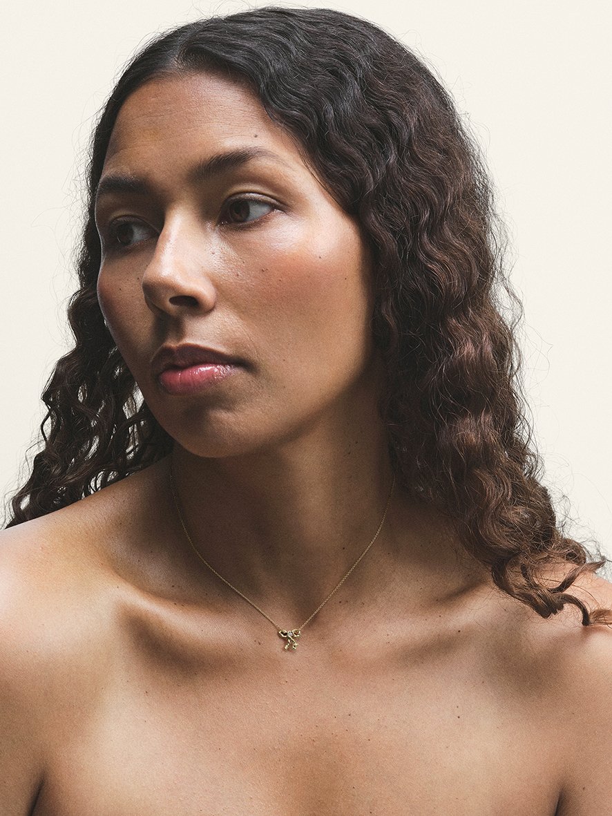A close-up of a woman with curly brown hair wearing a gold necklace with a bow pendant, looking to the side against a plain background.