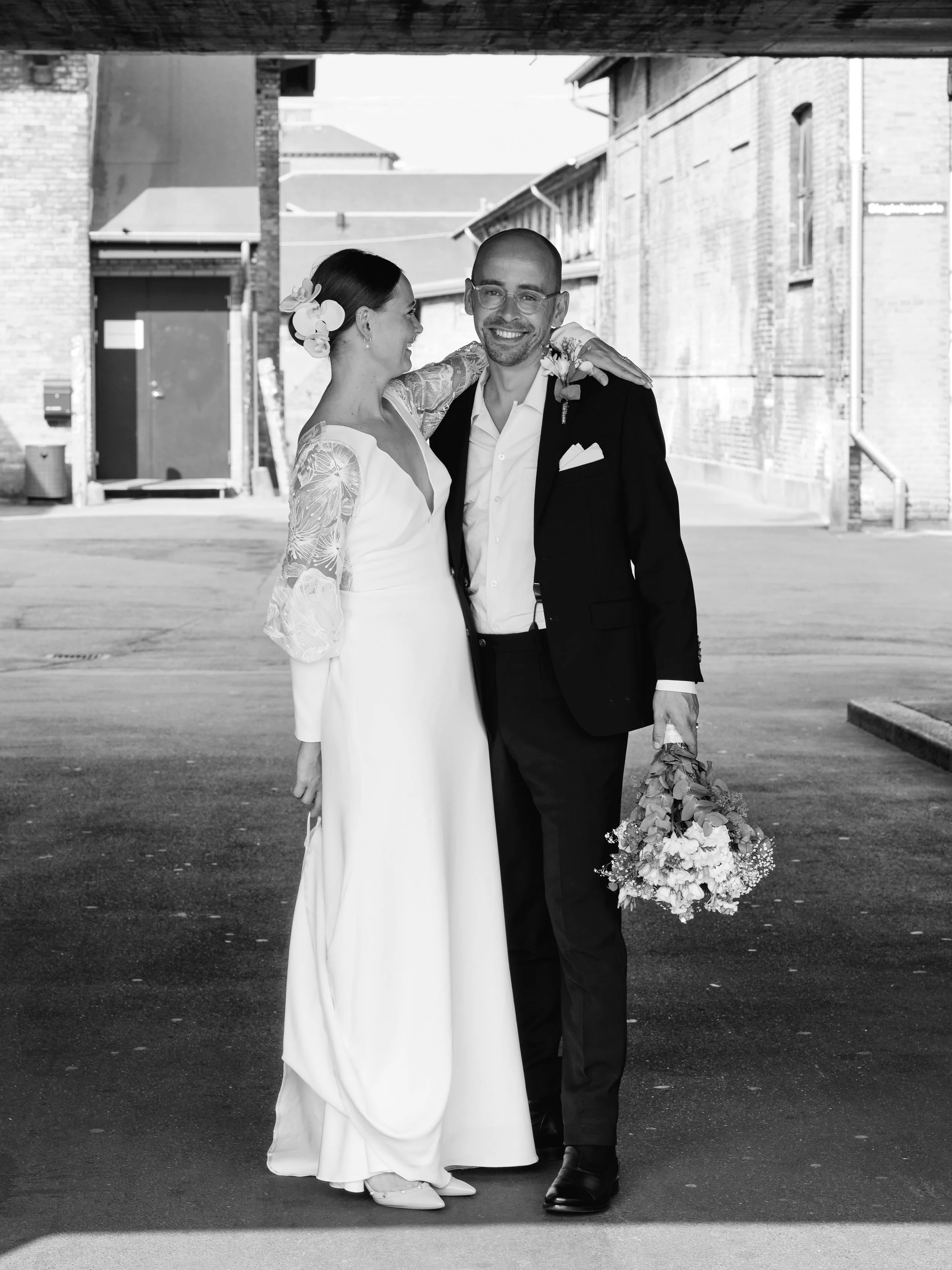 A black and white photo of a bride and groom standing outdoors. The bride is wearing a long white dress with lace sleeves and flowers in her hair. The groom is in a dark suit, white shirt, and holding a bouquet of flowers. They are smiling and sharing an affectionate moment.
