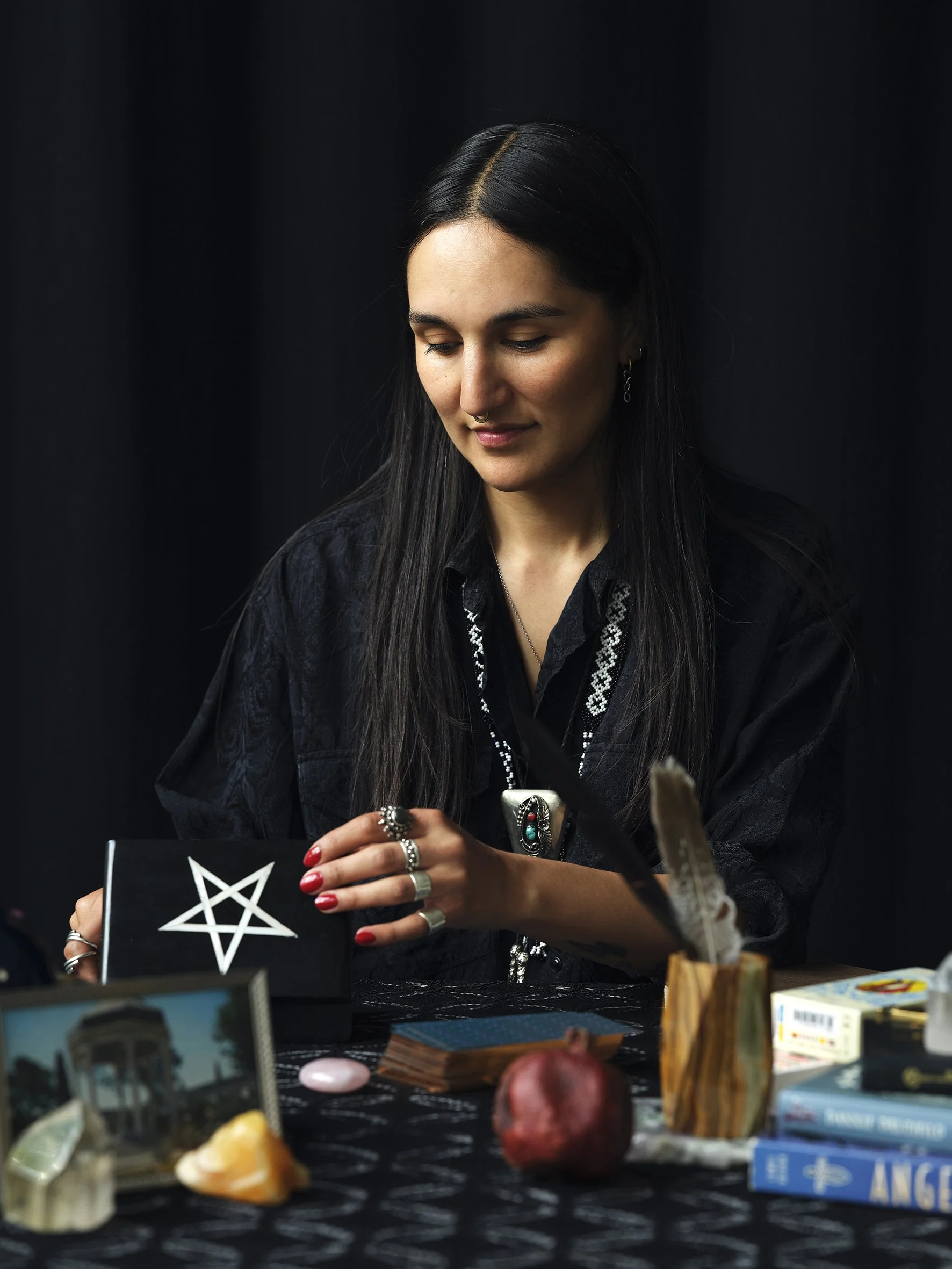 A woman with long black hair dressed in black, with jewelry, is sitting at a table with various objects including tarot cards, a pomegranate, and decorative items, holding a black box with a white pentagram symbol.