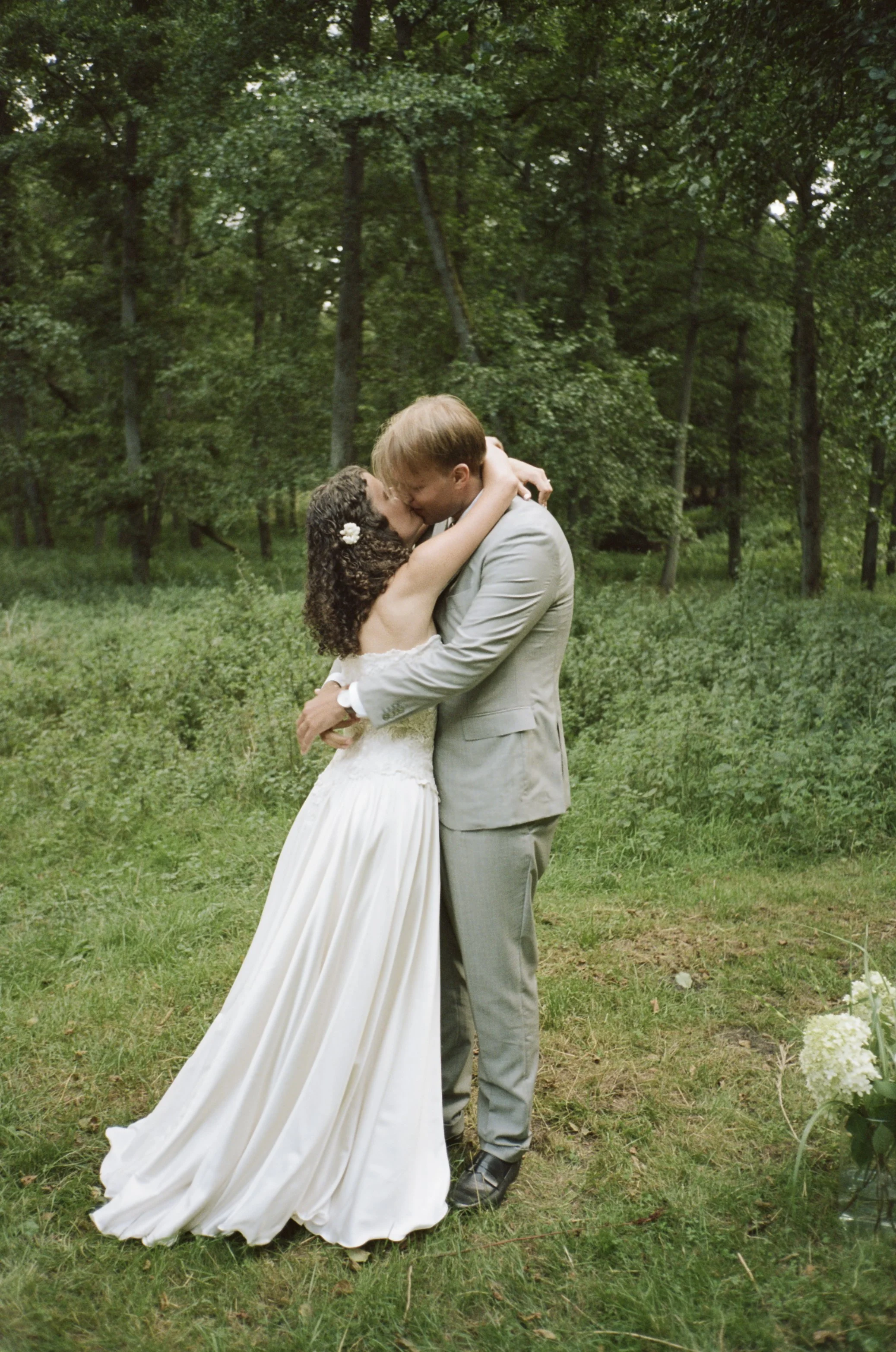 Couple in wedding attire kissing in a green outdoor setting with trees and grass.