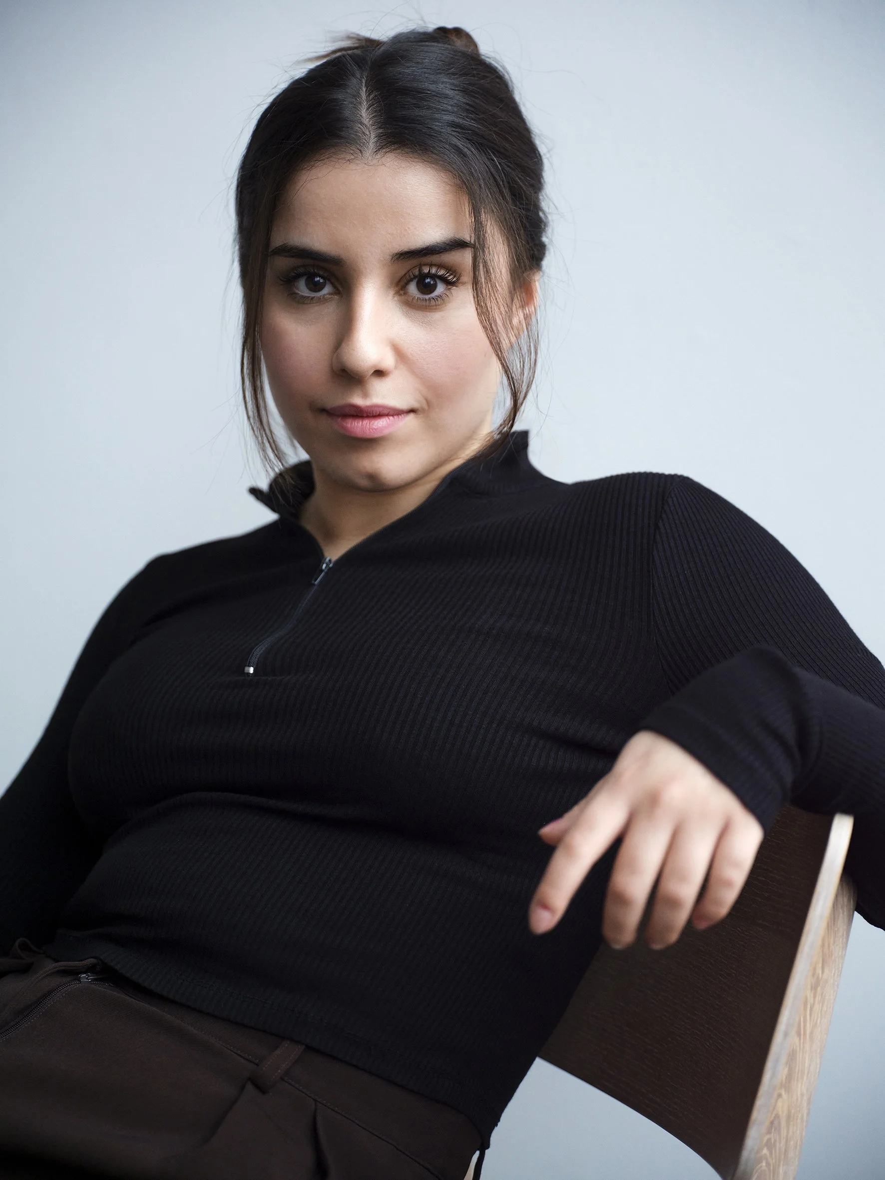 A woman sitting on a wooden chair, wearing a black long-sleeve zip-up shirt, looking confidently at the camera.