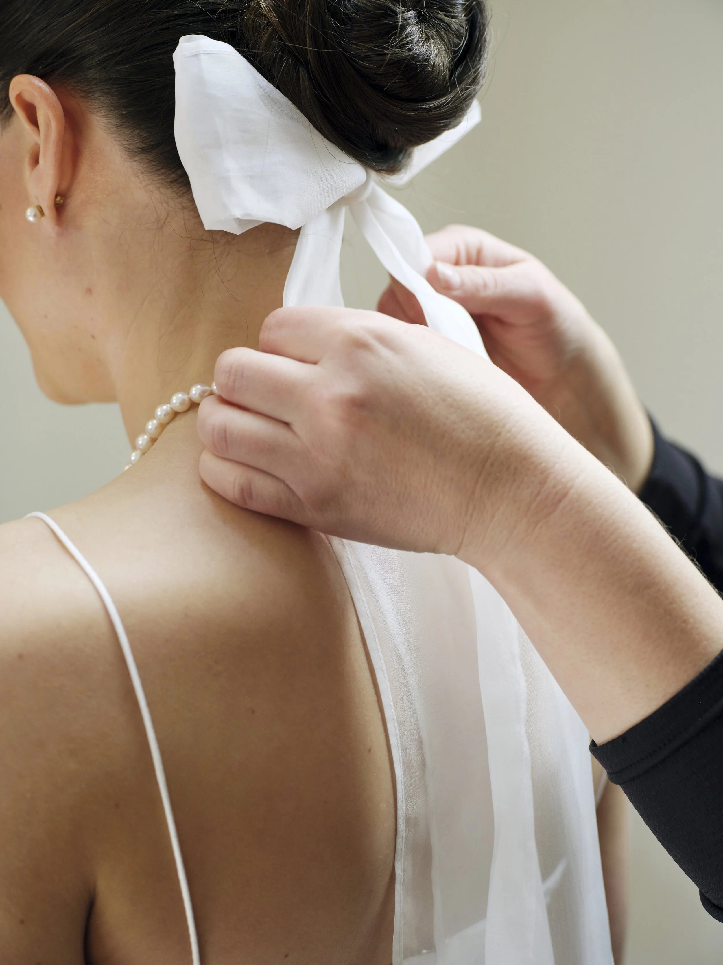 A woman with dark hair styled in an updo, wearing pearl earrings and a pearl necklace, is having a white ribbon tied around her neck by another person.