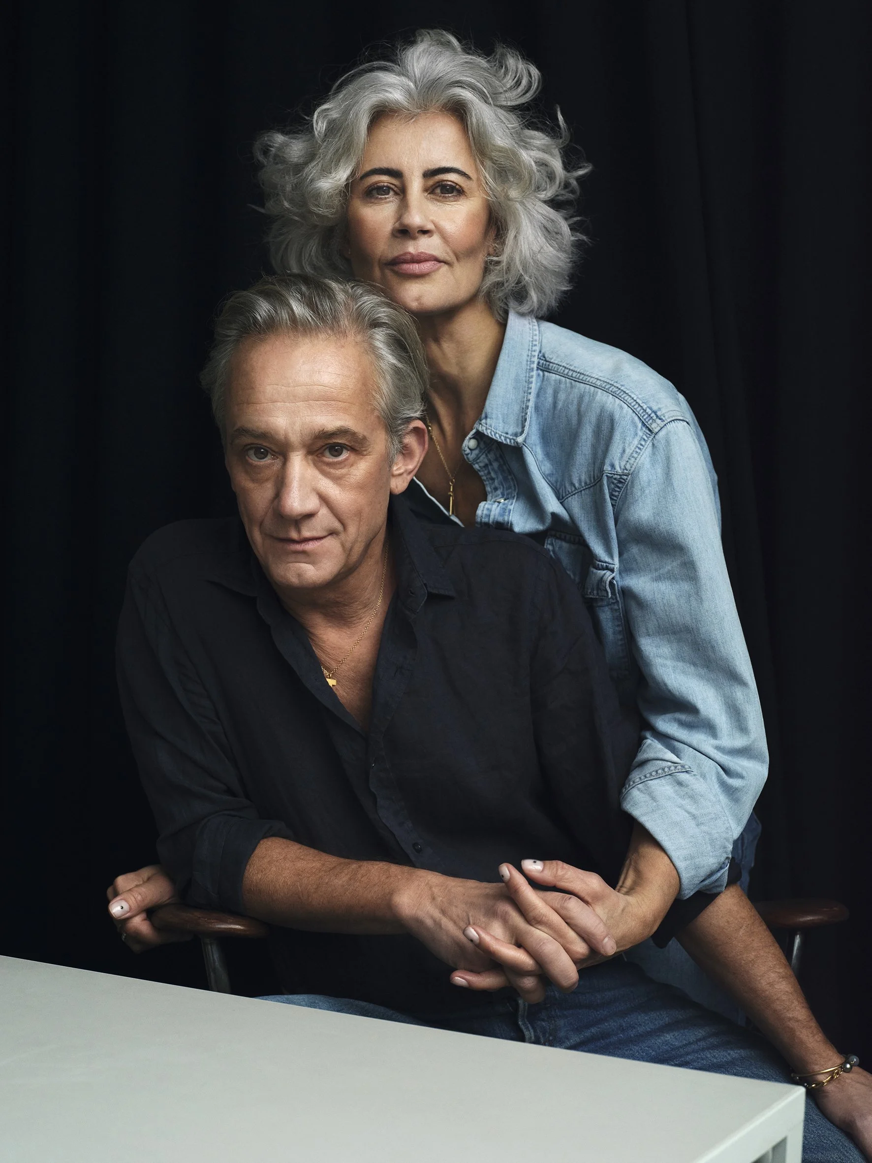 A colour portrait of a mature woman with curly gray hair standing behind an man with gray hair, sitting at a table, both looking at the camera against a dark background. couples photography in vouge expensive feeling