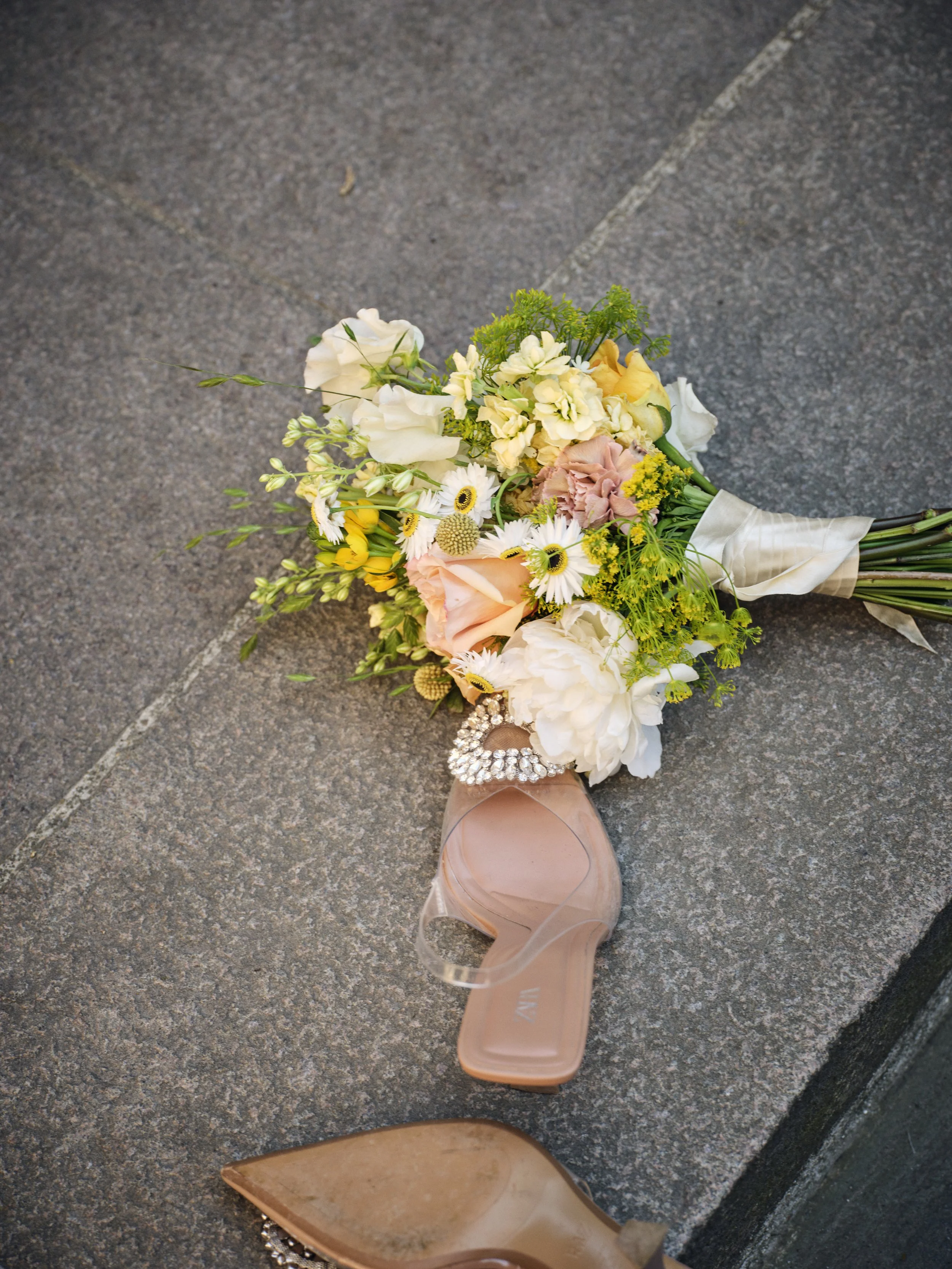 A bridal bouquet with pink, white, and yellow flowers resting on a concrete sidewalk alongside a women's high-heeled shoe with a decorative rhinestone ankle strap and pointed toe.