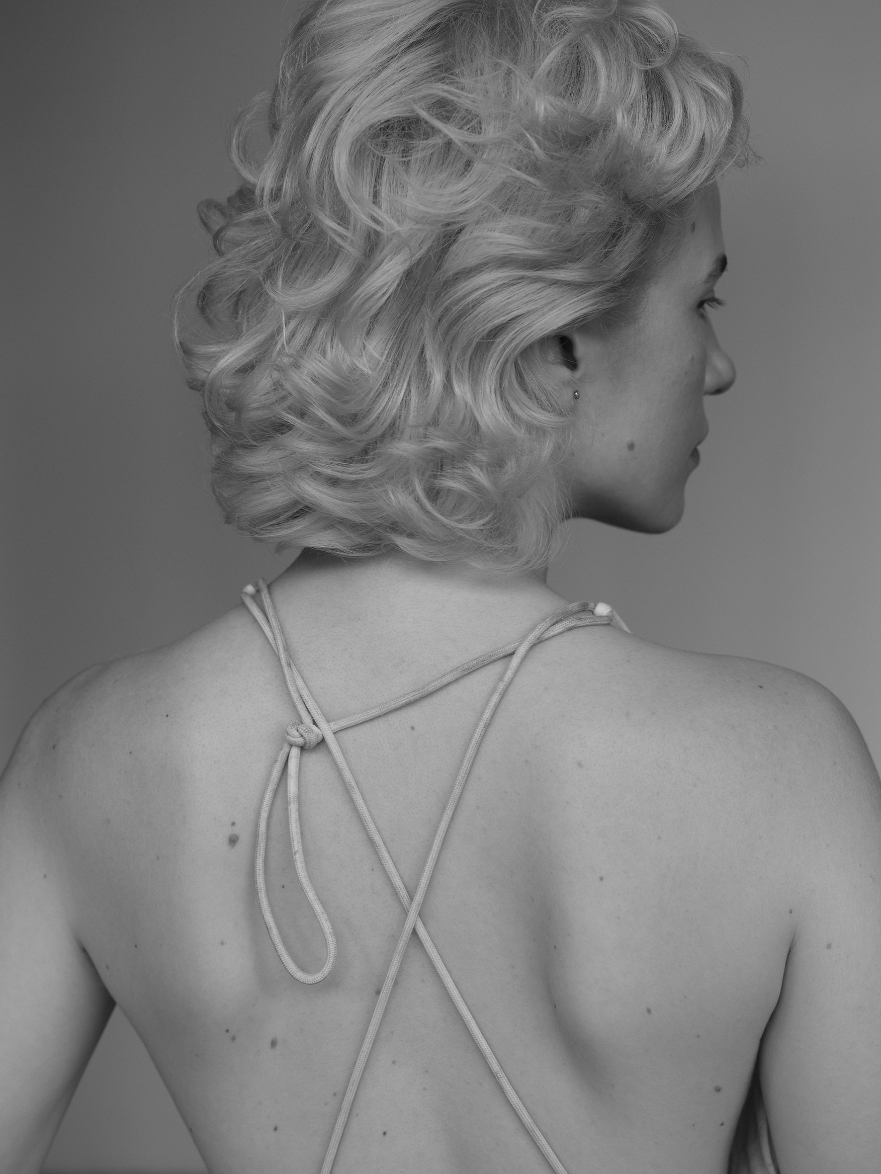 A black and white photo of a woman with short, curly blonde hair, seen from the back, wearing a thin-strapped top.