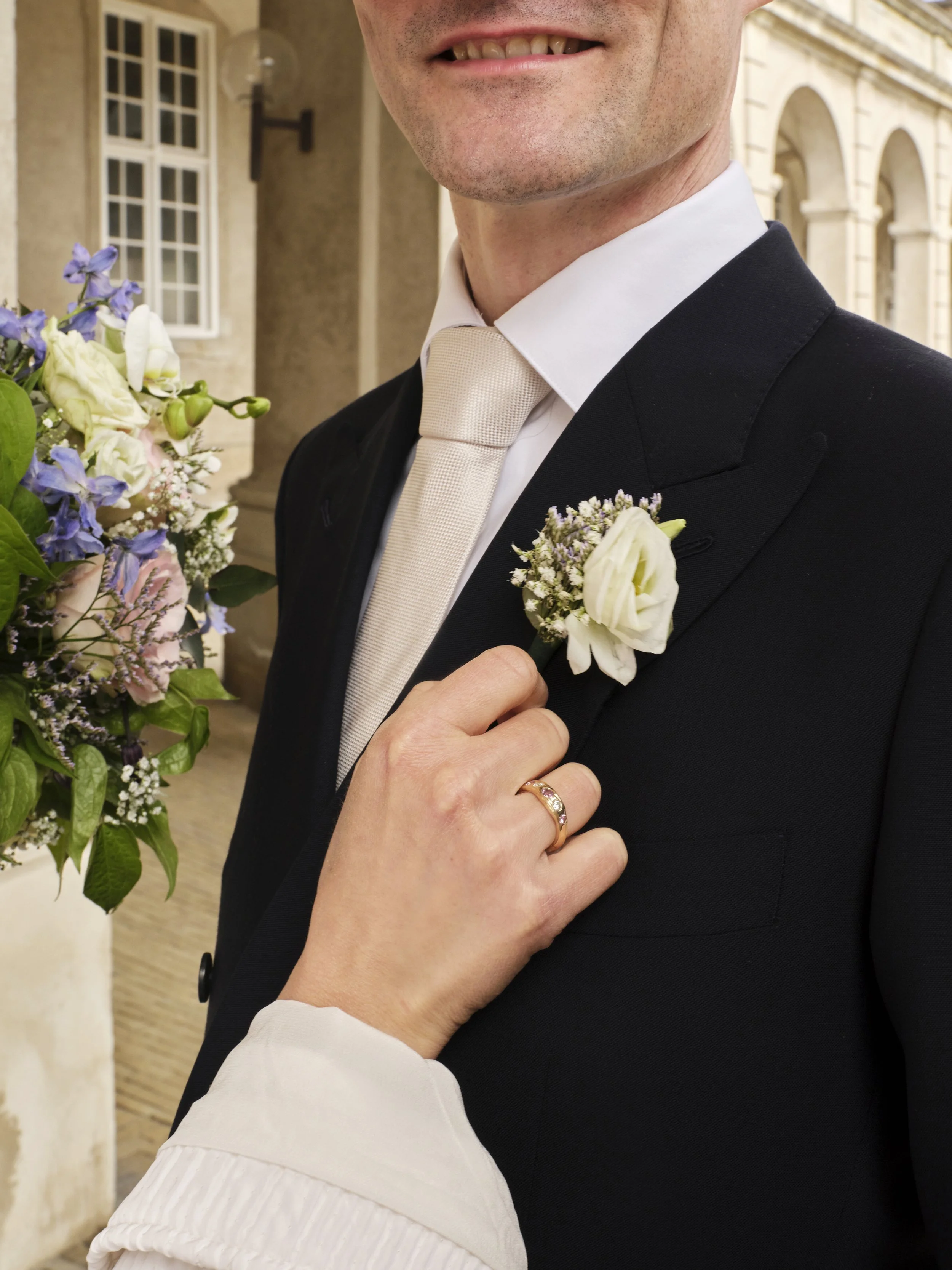 Close-up of a groom in a black suit with a light-colored tie and a boutonniere, and a hand with a ring adjusting the boutonniere.