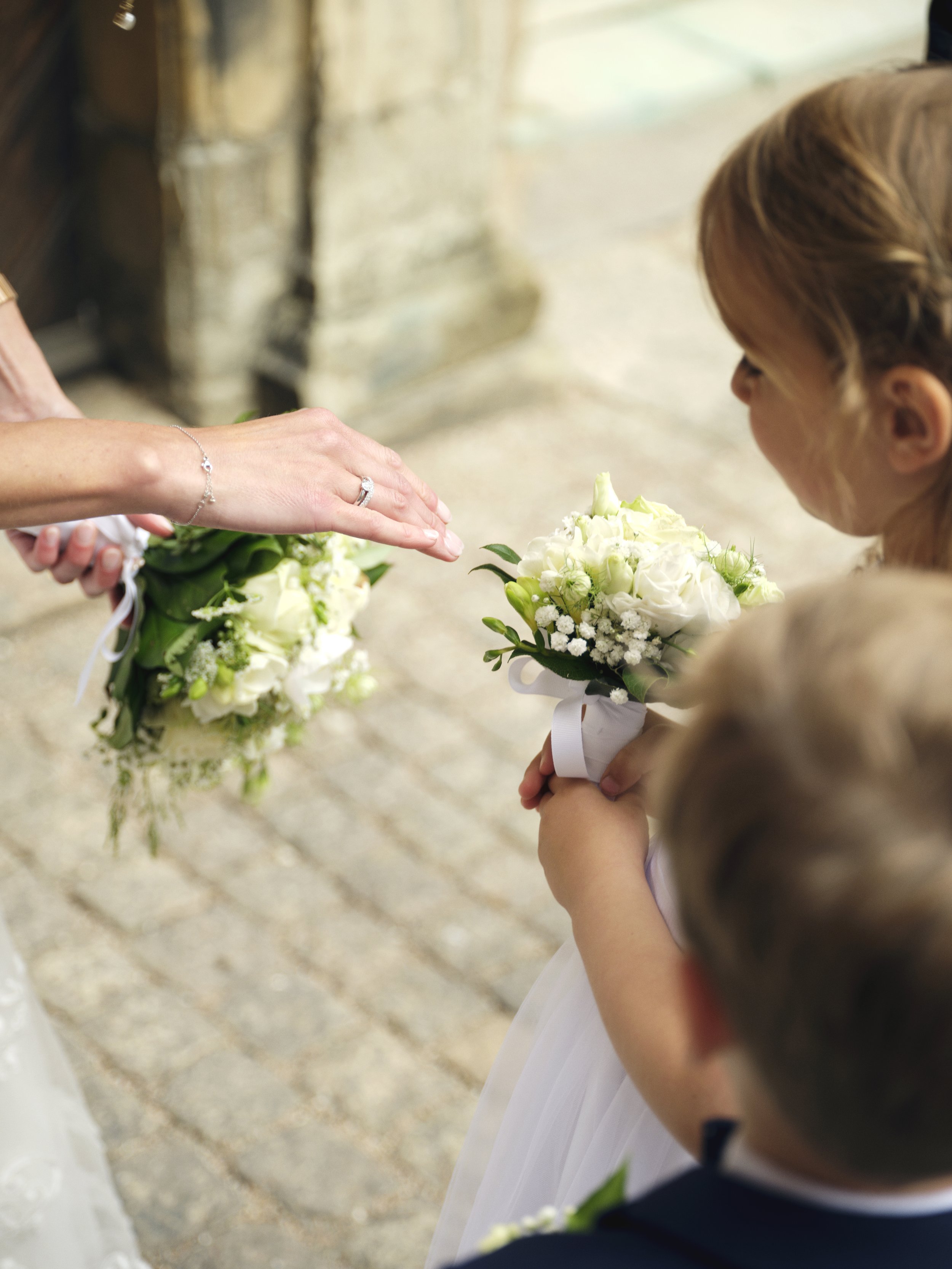 A woman holding a bouquet of white flowers is about to give them to a young girl during a wedding ceremony. The woman is wearing a ring and a bracelet, and the girl has a white dress with a bow. Another child, a boy, is visible in the foreground, all standing outdoors on a stone-paved surface.