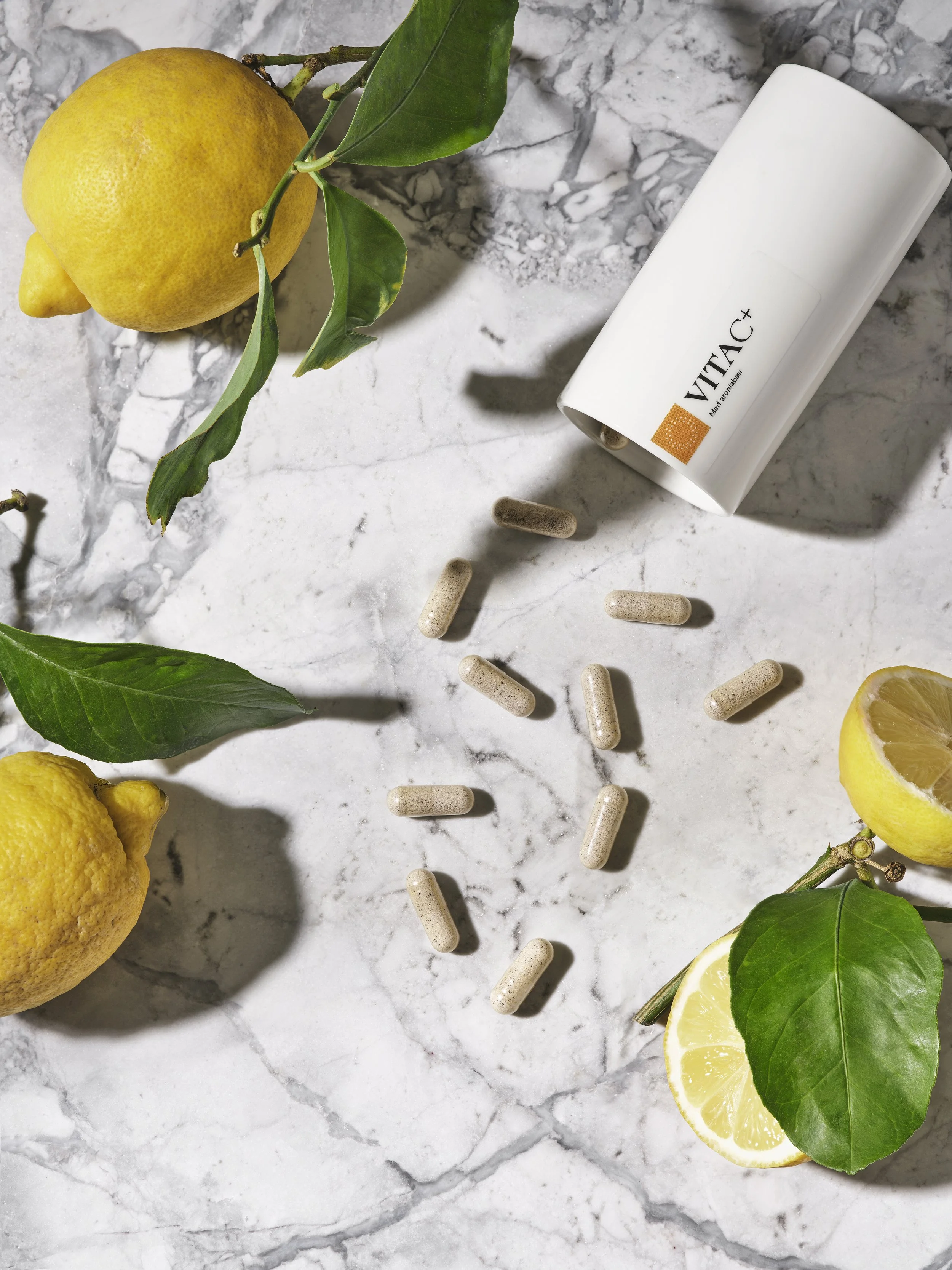 Lemon fruits with green leaves, a white bottle labeled VITA+, and scattered beige capsules on a white marble surface.