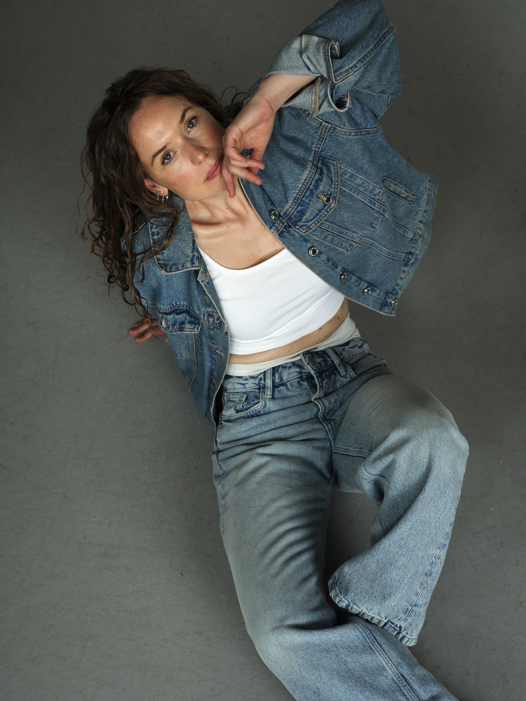 A woman with curly hair in casual blue jeans and a denim jacket, lying on the floor, looking up at the camera with her hand near her face. actor portait.