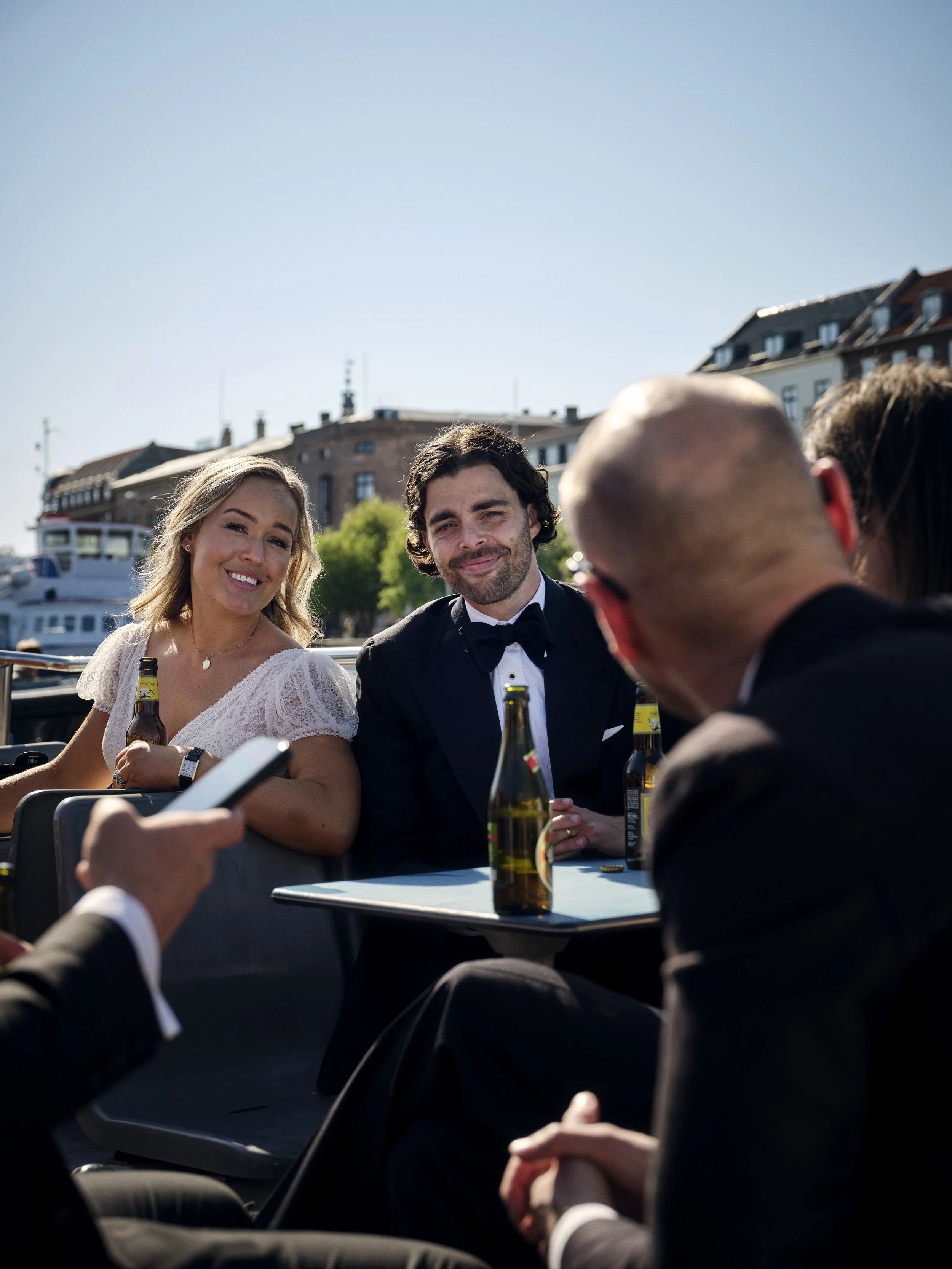 People in formal attire enjoying drinks at an outdoor gathering near a waterfront, with boats and buildings in the background on a sunny day.