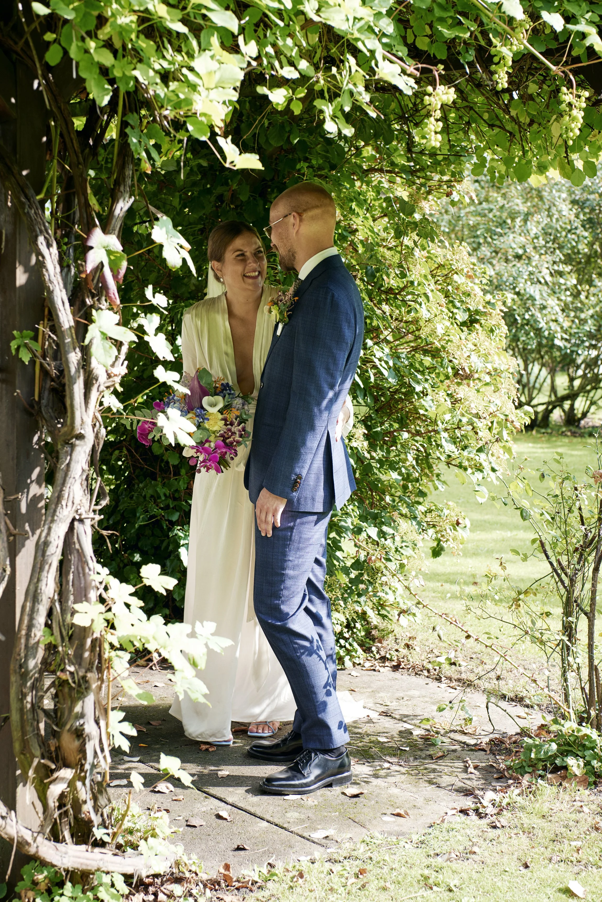 A bride and groom stand close together outdoors, surrounded by green foliage, sharing an intimate moment on their wedding day.