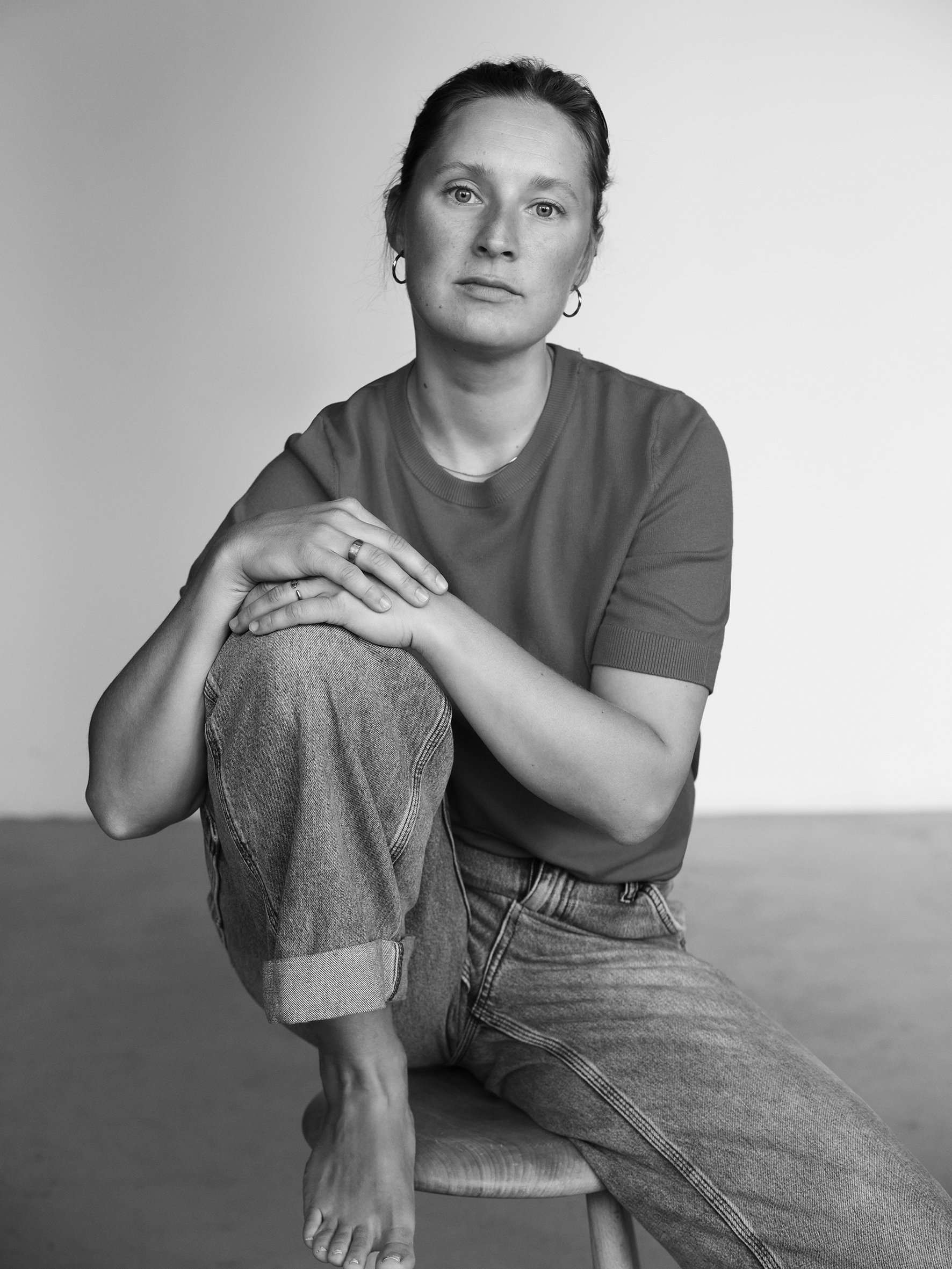 Black and white portrait of a woman sitting on a stool with her left knee raised, wearing a t-shirt and jeans, looking directly at the camera. actor portrait Anna