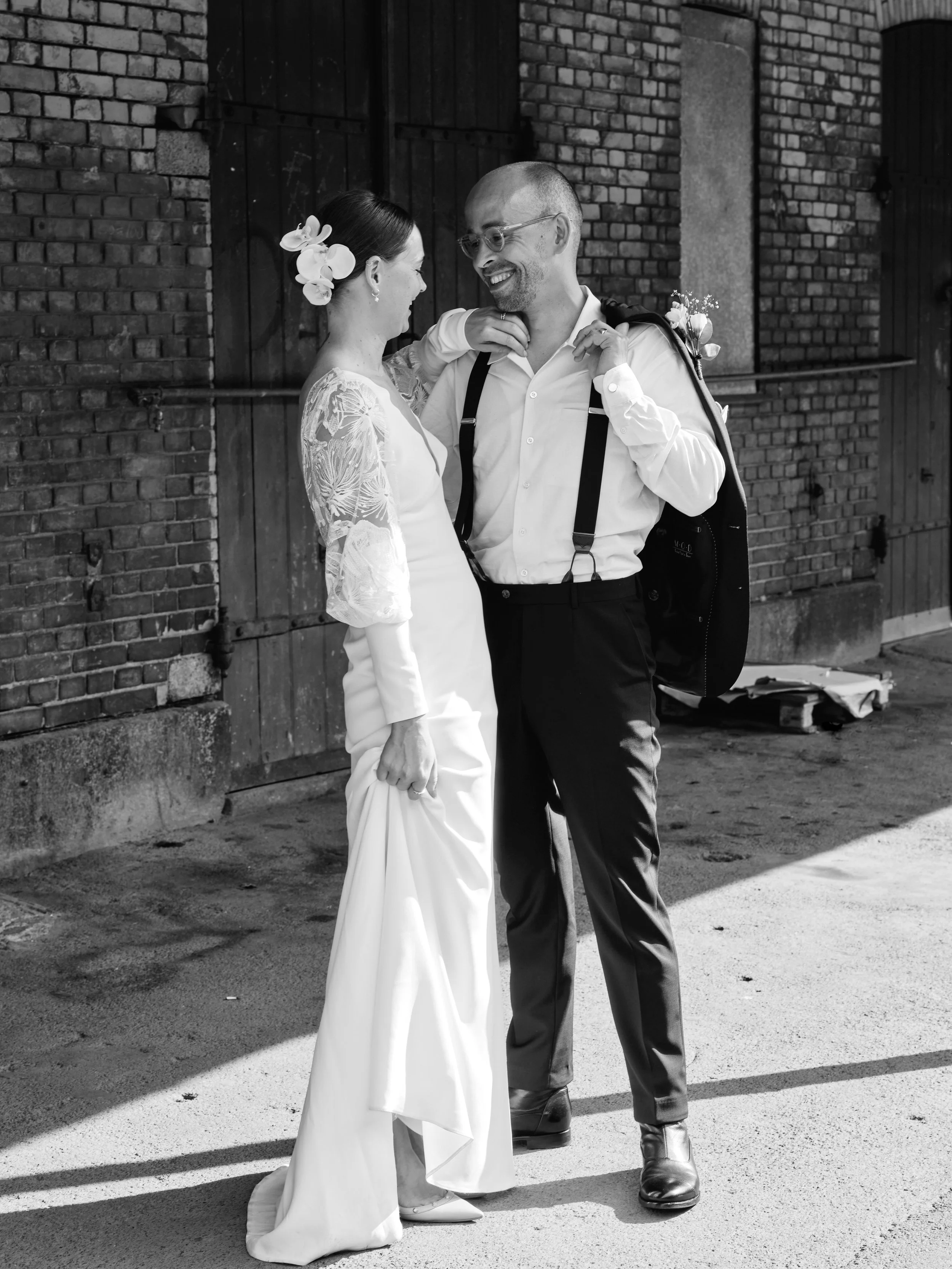 A black and white photo of a bride and groom standing close, smiling at each other. The bride is in a long white dress with lace sleeves, holding her dress with one hand, and has flowers in her hair. The groom is wearing a white shirt, dark pants, suspenders, glasses, and carries a backpack. They are outdoors in front of a brick and wooden wall.