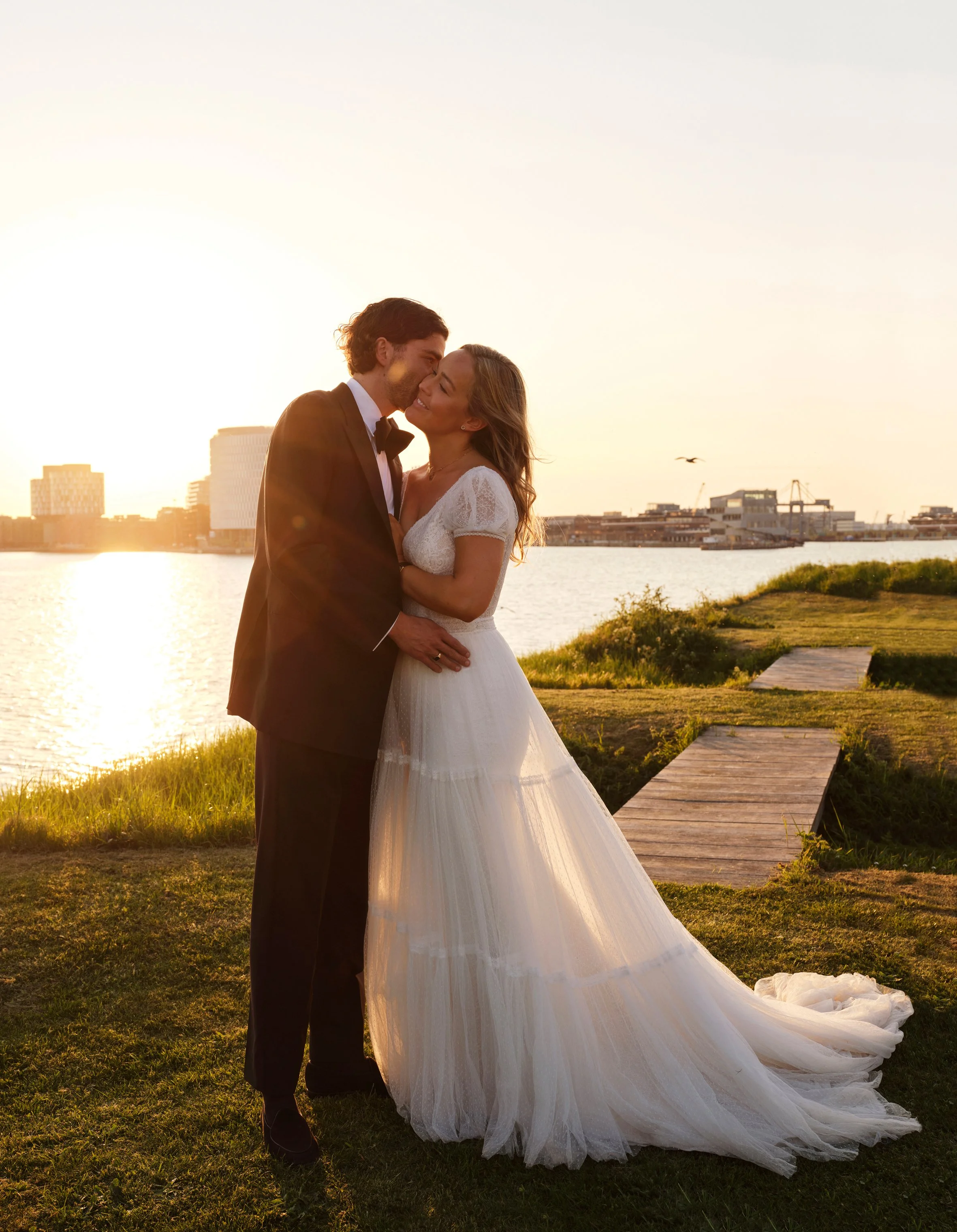 A newlywed couple in wedding attire embracing by a river at sunset, with city buildings in the background.