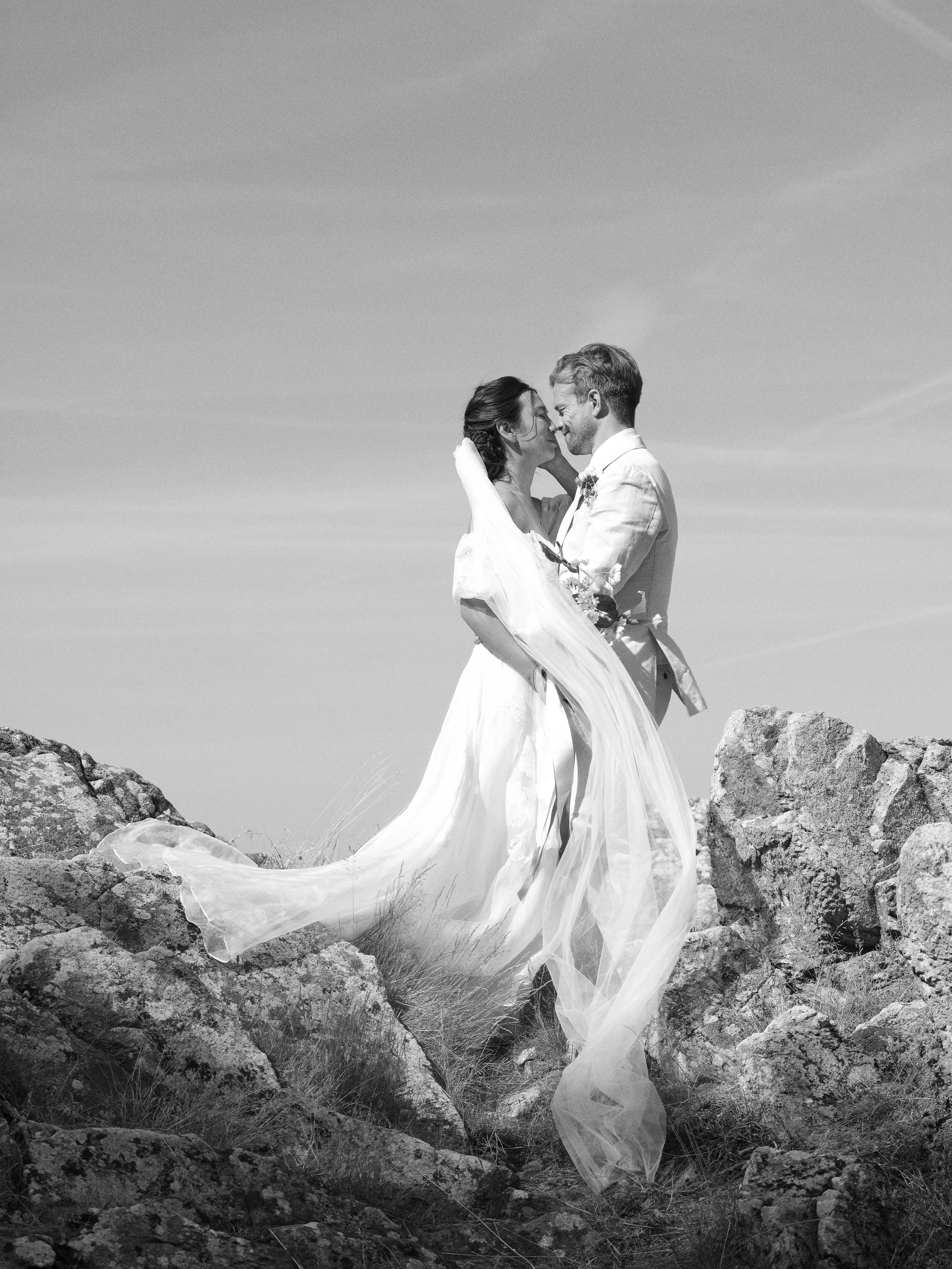 Black and white photo of a bride and groom standing on rocks, embracing and touching noses, outdoors with a clear sky in the background.