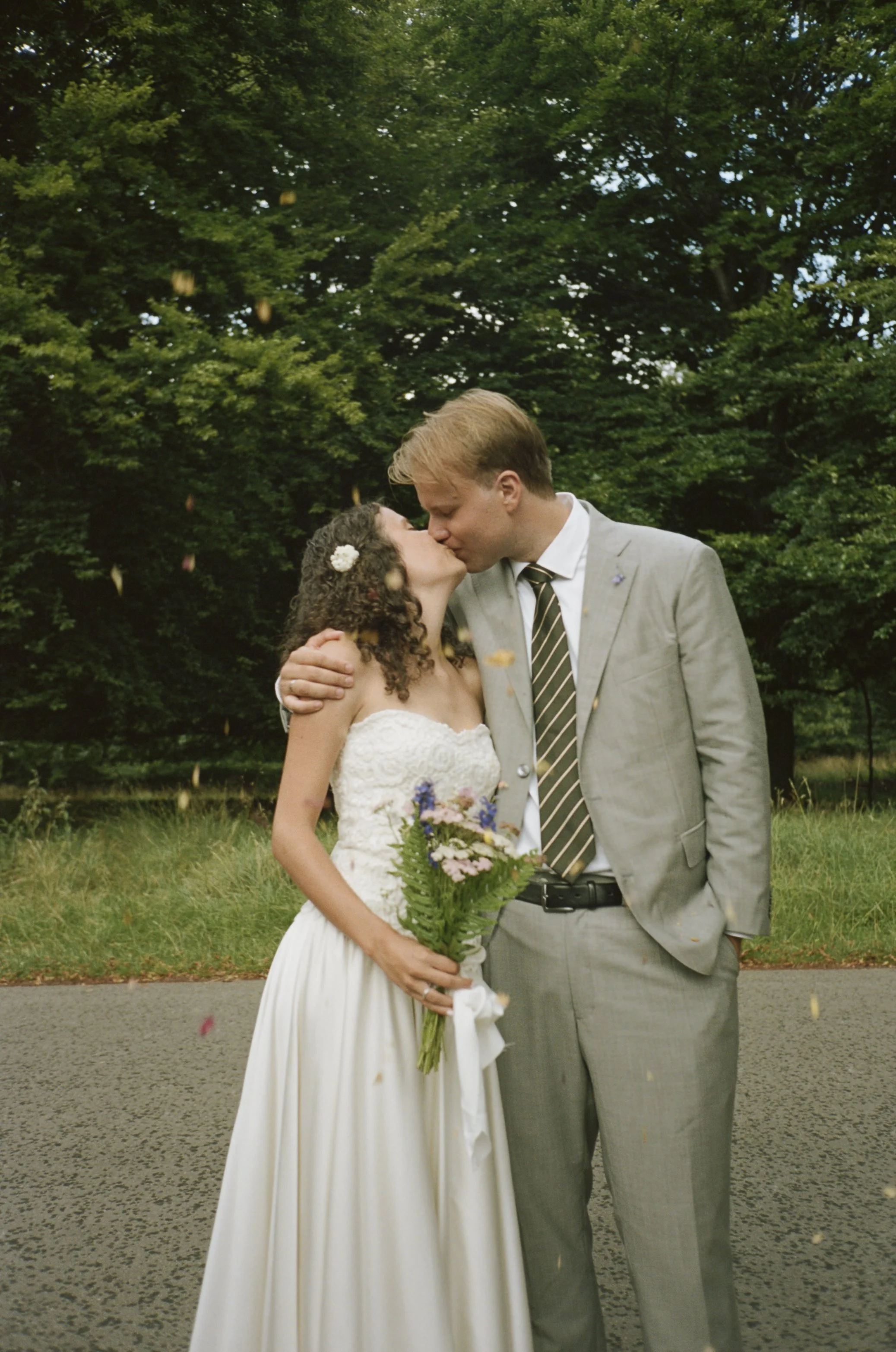 A bride and groom sharing a kiss outdoors, with the bride holding a bouquet of flowers, surrounded by green trees and falling leaves or petals.