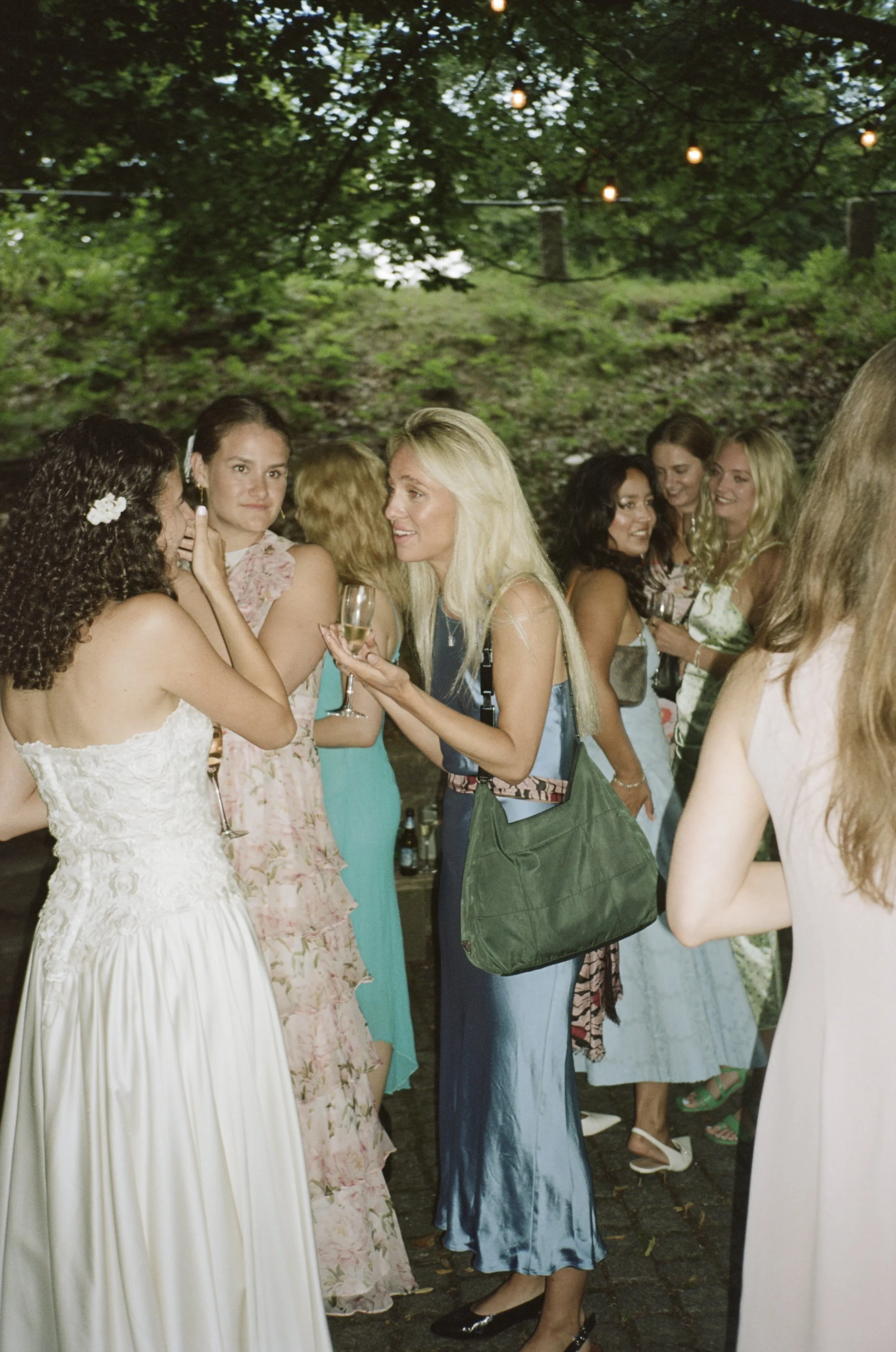 Group of women socializing outdoors at a party, some holding drinks, under string lights in a green, wooded setting.