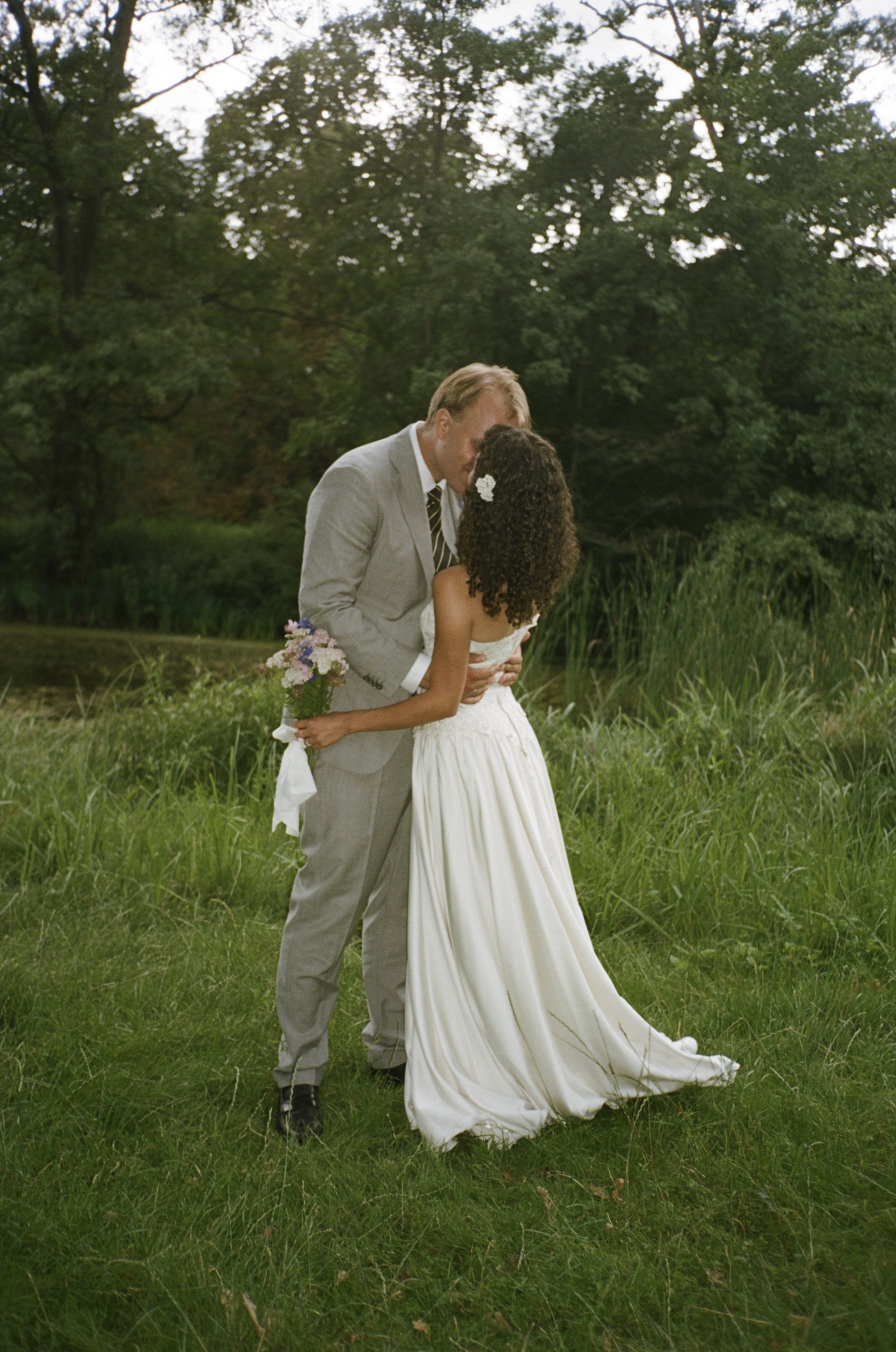 A couple sharing a kiss outdoors by a pond, with the woman holding a bouquet of flowers and wearing a white dress.