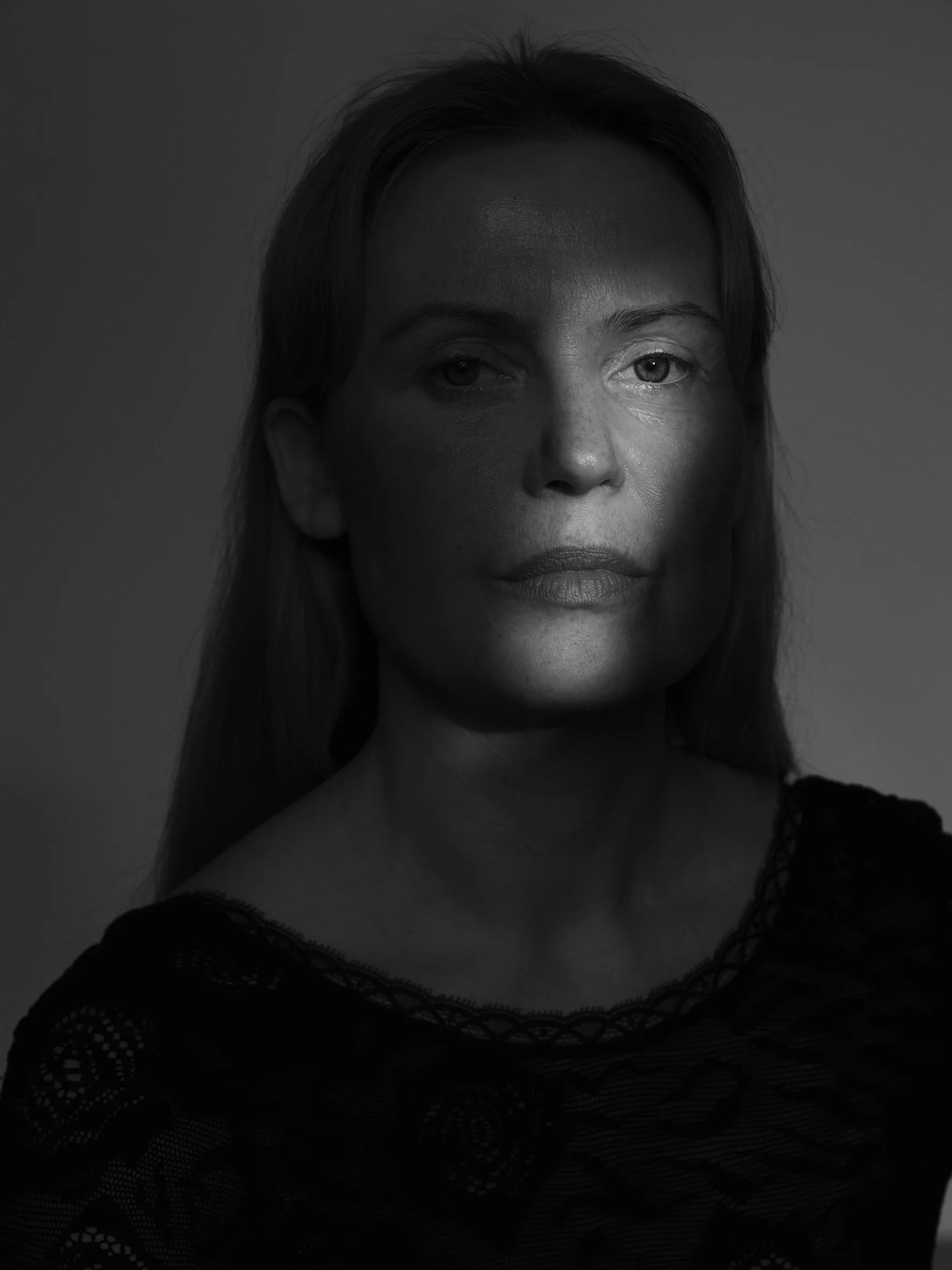 Black and white portrait of a woman with long-length hair, looking directly at the camera with a neutral expression. shadow and light combination showing the detail on eye and lip of actor and scripter Trine Rosa Bunde.