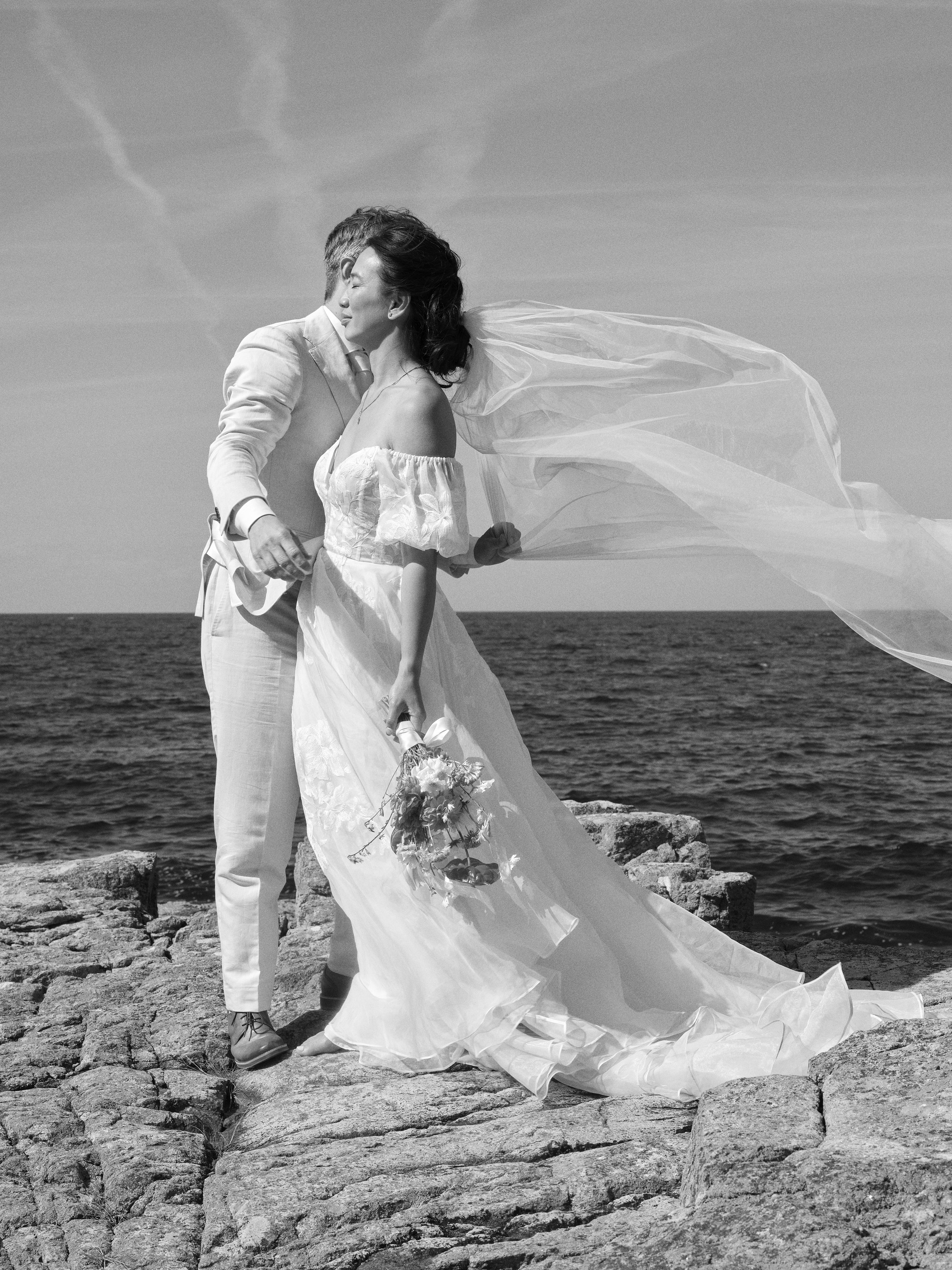 A black and white photo of a bride and groom standing on rocks by the sea, with the bride holding a bouquet and her veil flowing in the wind, wearing wedding attire.
