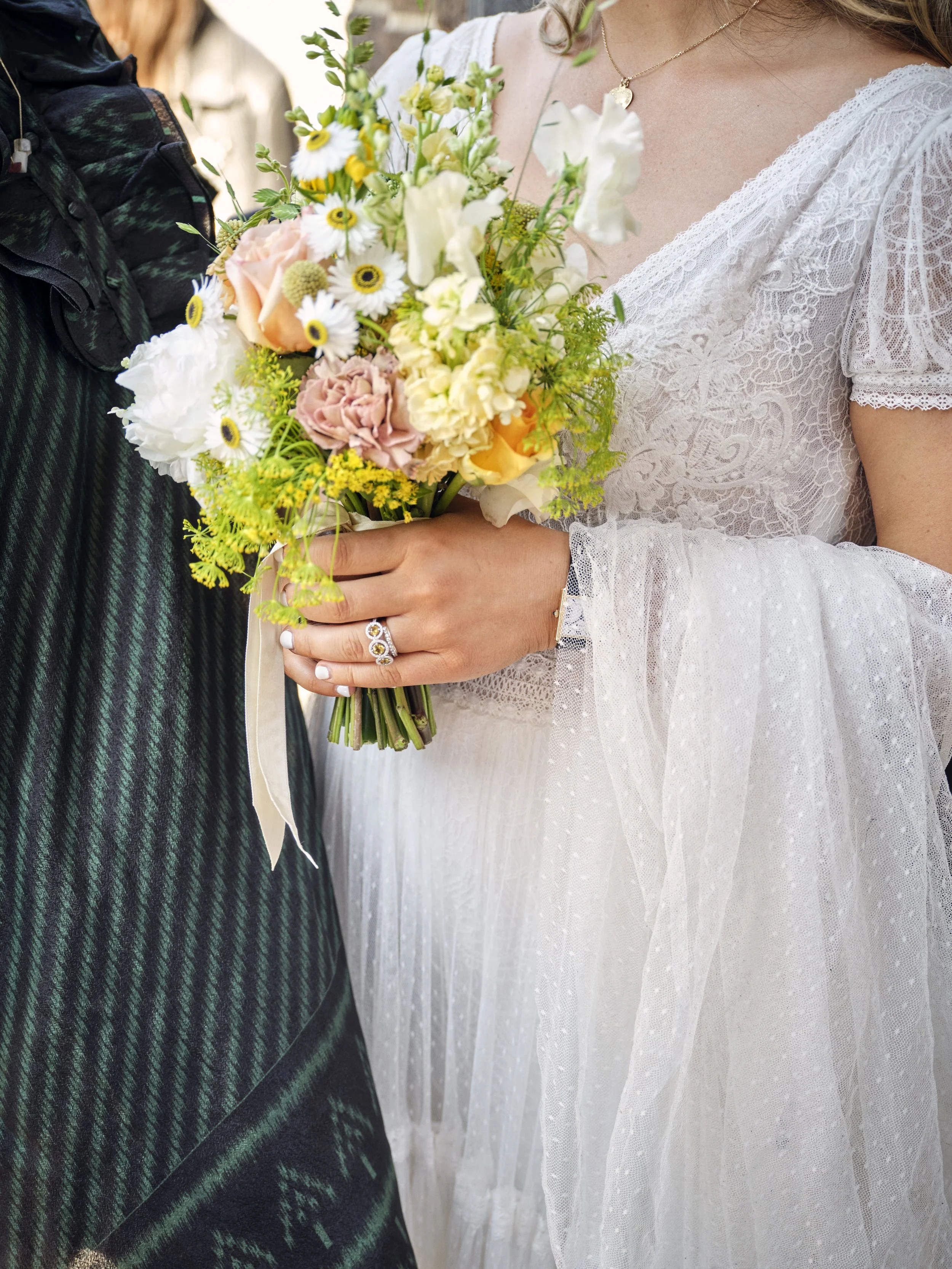 Close-up of a woman in a white lace dress holding a bouquet of various flowers, including roses, daisies, and greenery, with a ring on her finger and a gold necklace.