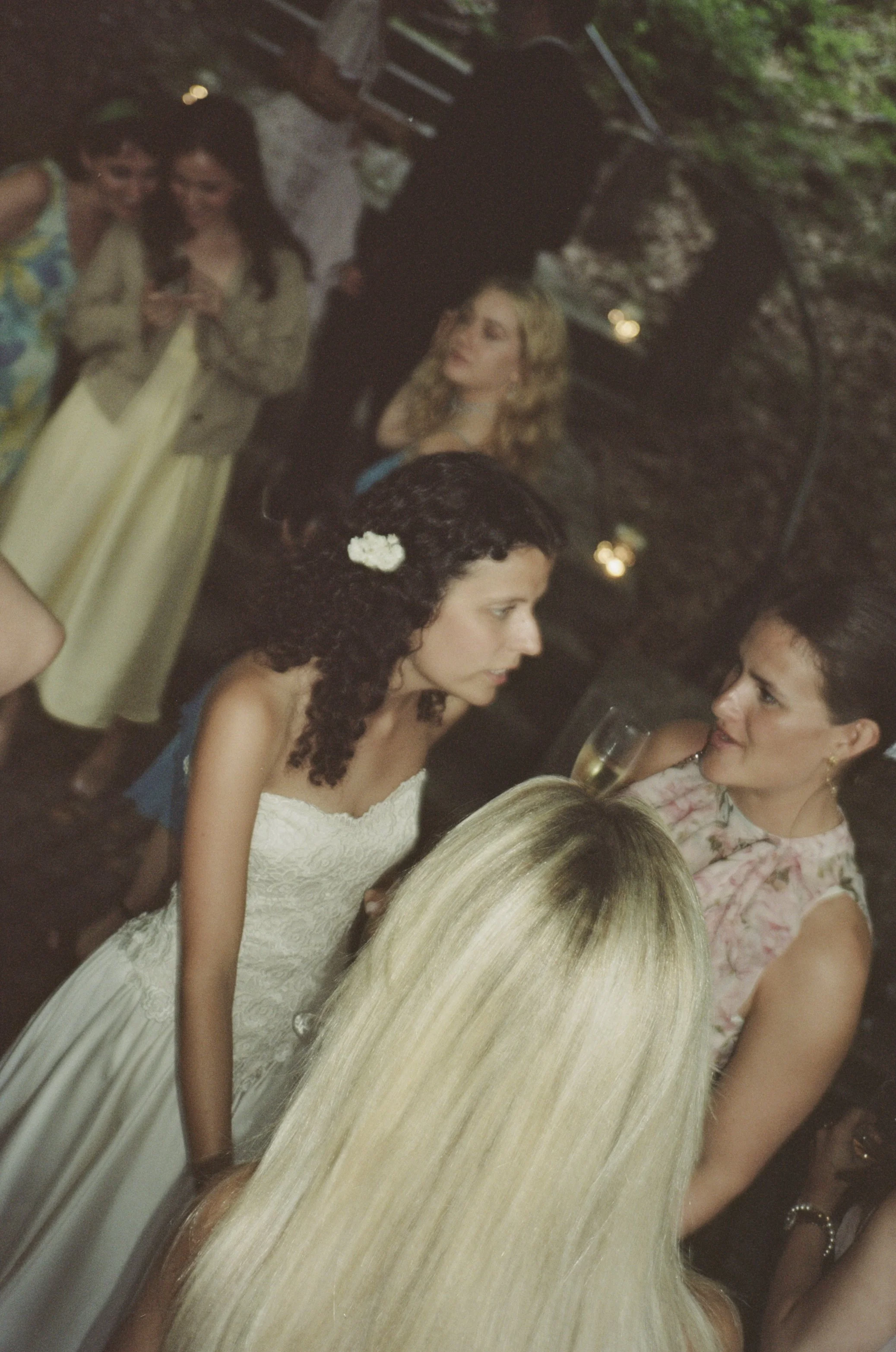 A group of women at a social gathering, with one wearing a white lace dress and a flower in her dark curly hair, engaging in conversation, with other women in the background and some holding drinks.