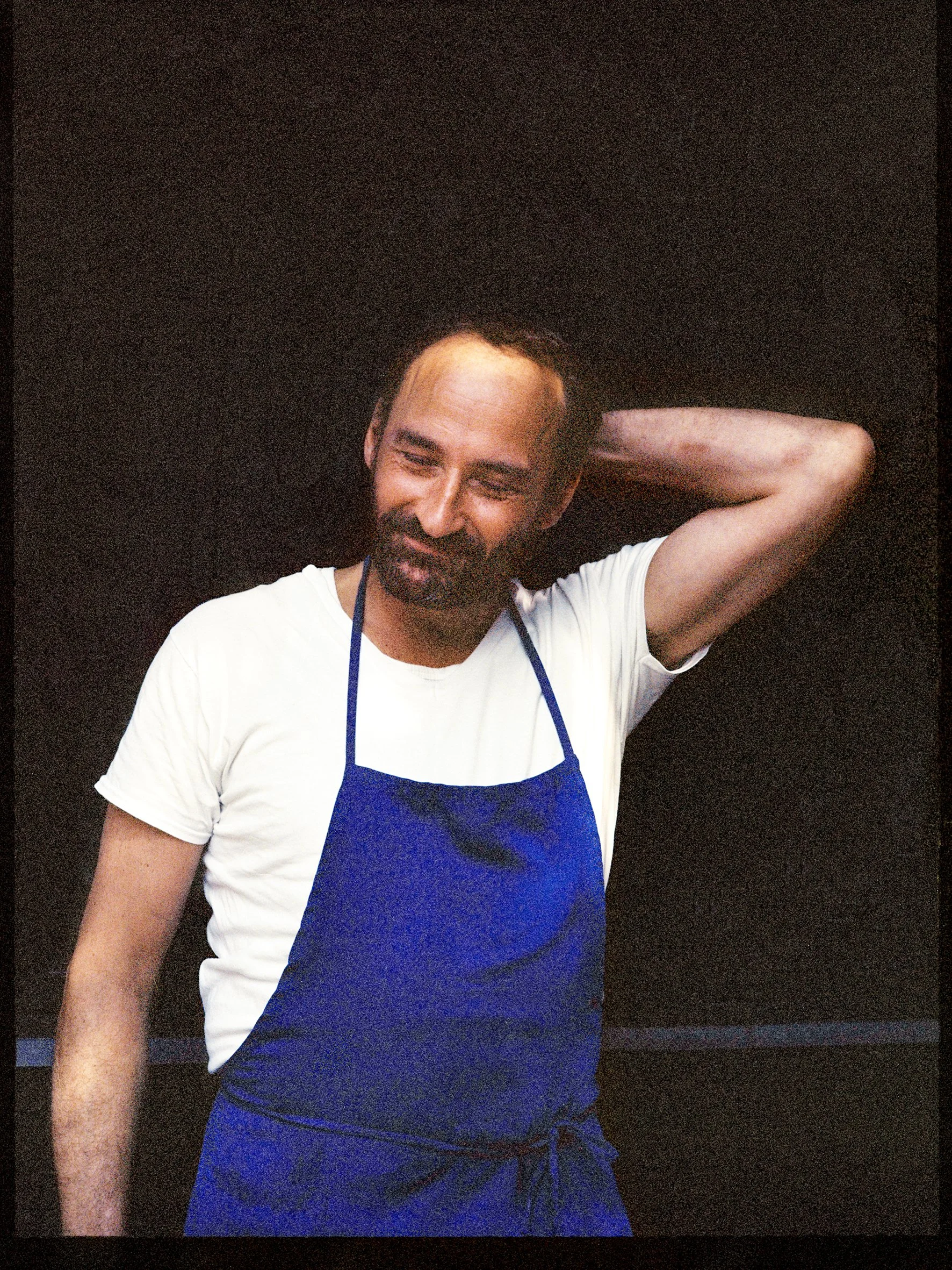A man with a beard and mustache, wearing a white t-shirt and a blue apron, standing against a dark background. He has one arm behind his head and is smiling with his eyes closed.