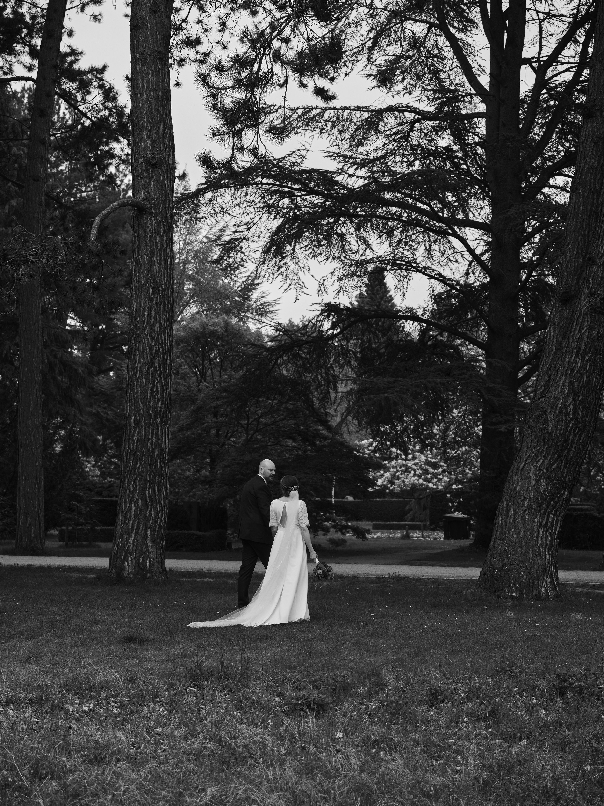 A black and white photo of a bride and groom walking in a park, surrounded by tall trees, with the bride holding a bouquet.