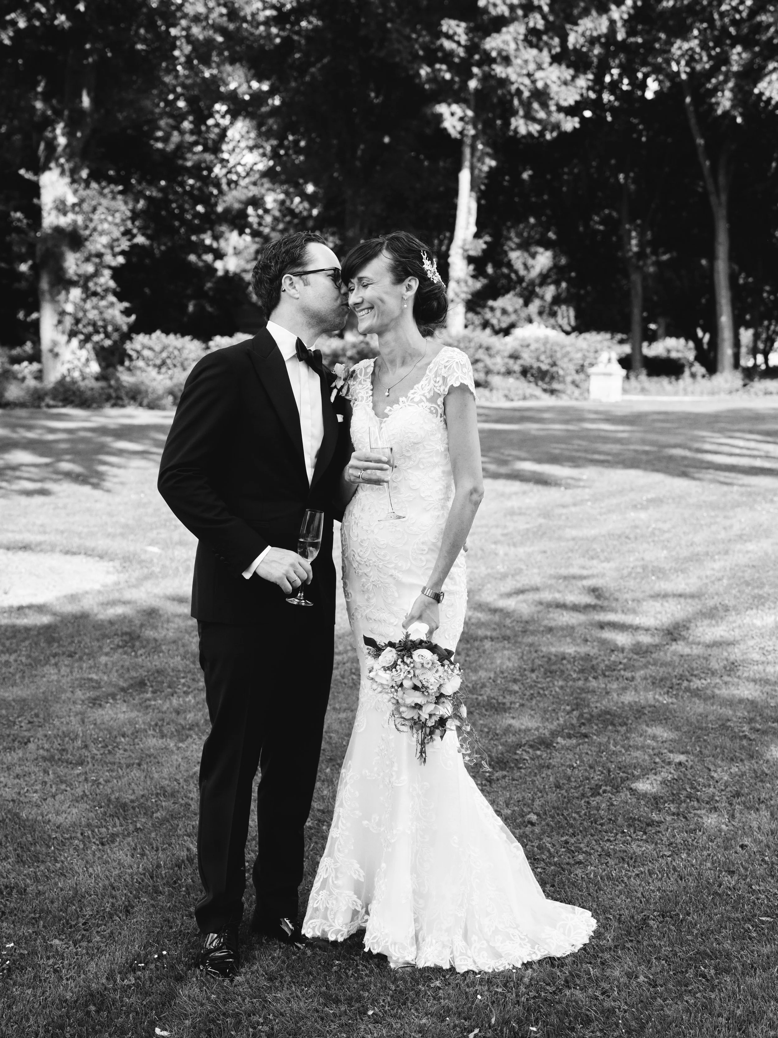 A newlywed couple in wedding attire sharing a kiss outdoors, both holding champagne glasses, with a woman holding a bouquet, in a grassy area with trees in the background.