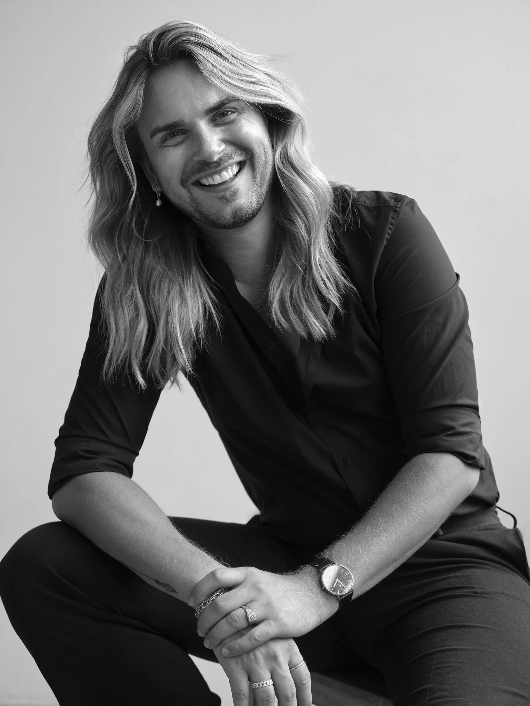 A smiling man with long wavy hair, wearing a dark shirt, sitting with one knee up, looking at the camera in a black-and-white portrait.