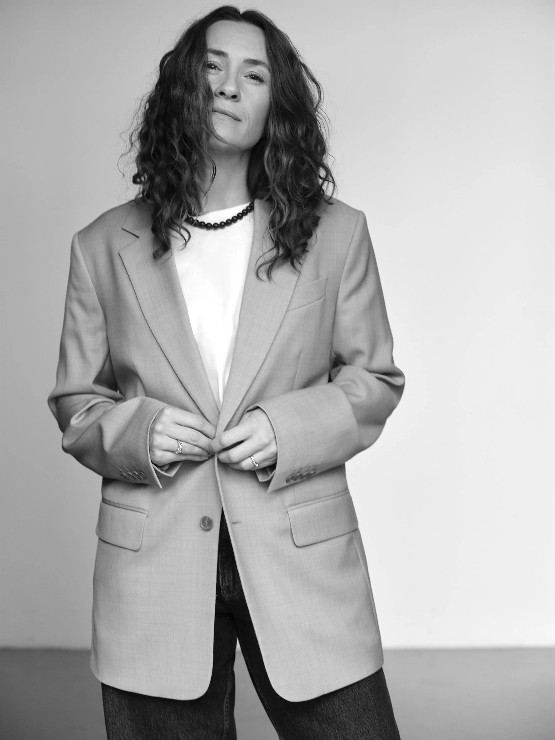 Black and white photo of a woman with wavy shoulder-length hair, wearing a blazer over a white shirt with a dark necklace, posing with a confident expression.