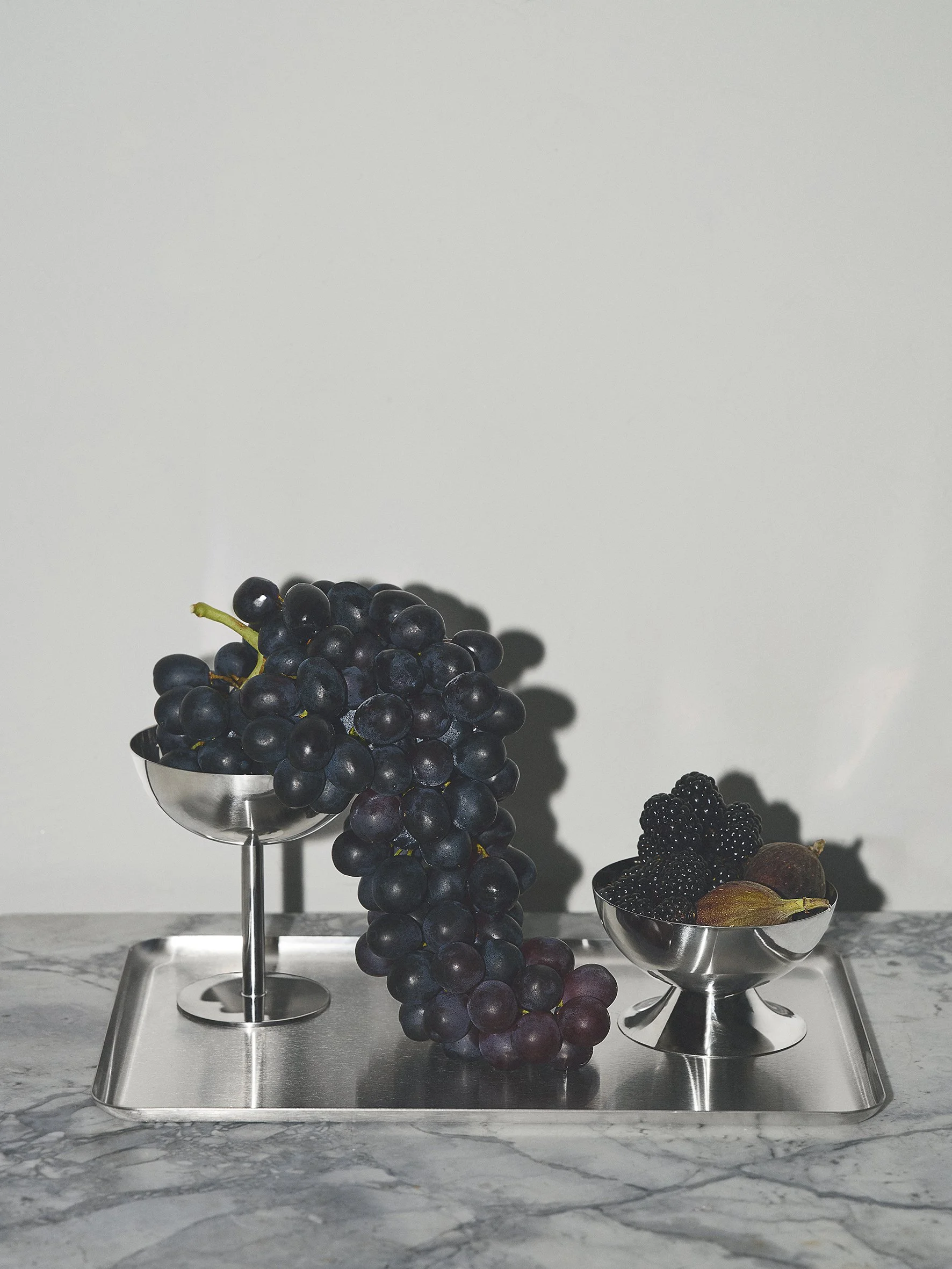 A stainless steel tray with two small metal bowls, one filled with black grapes and the other with blackberries and figs, on a marble surface against a plain wall. Fragmnt Copenhagen campaign with exclusive feelings. expensive and unique.