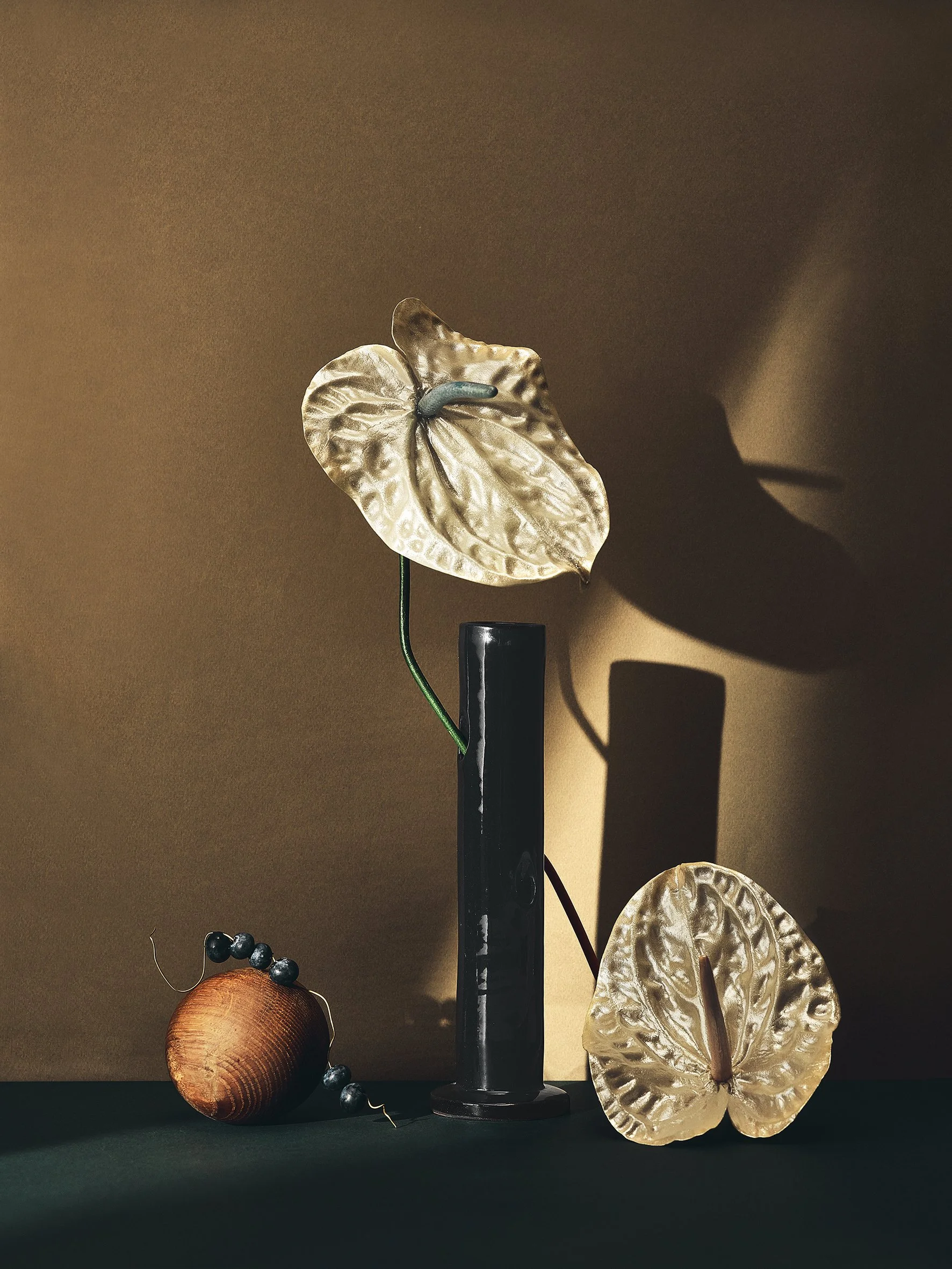 Decorative arrangement with a large metallic anthurium flower, a black cylindrical stand, a dried flower with a wooden bead, and a metallic anthurium flower shadow on a brown background. Reevein studio vase and flowes